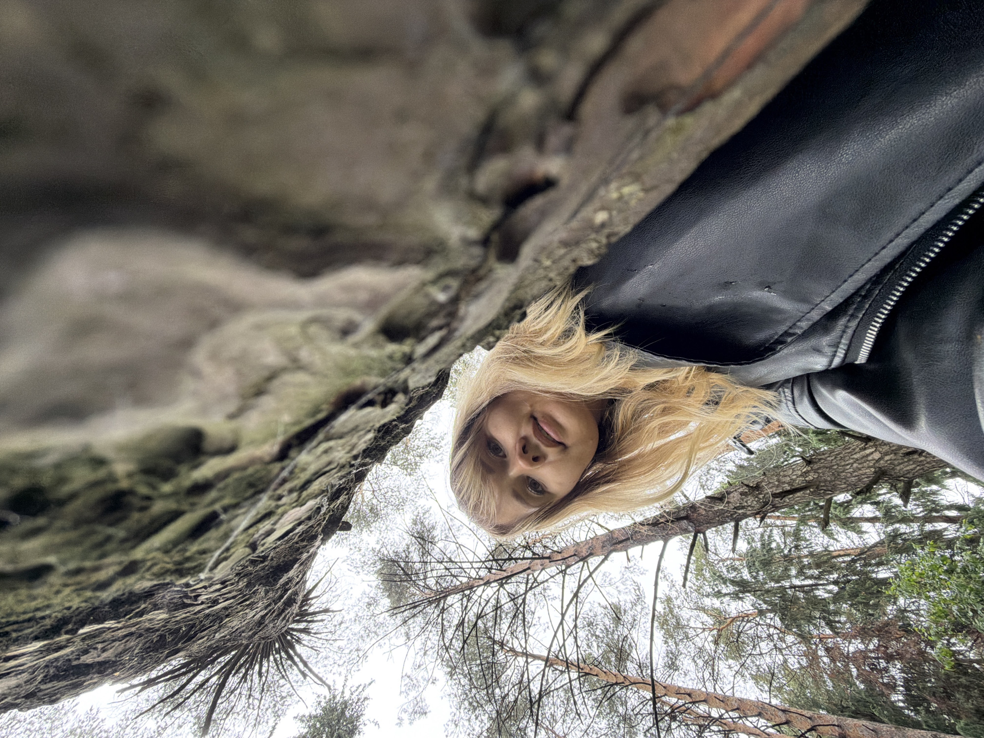 A woman with blonde hair is positioned under a rock formation, looking directly at the camera. She is wearing a black leather jacket. The backdrop consists of trees. The location is Chaikovsky, Russia.