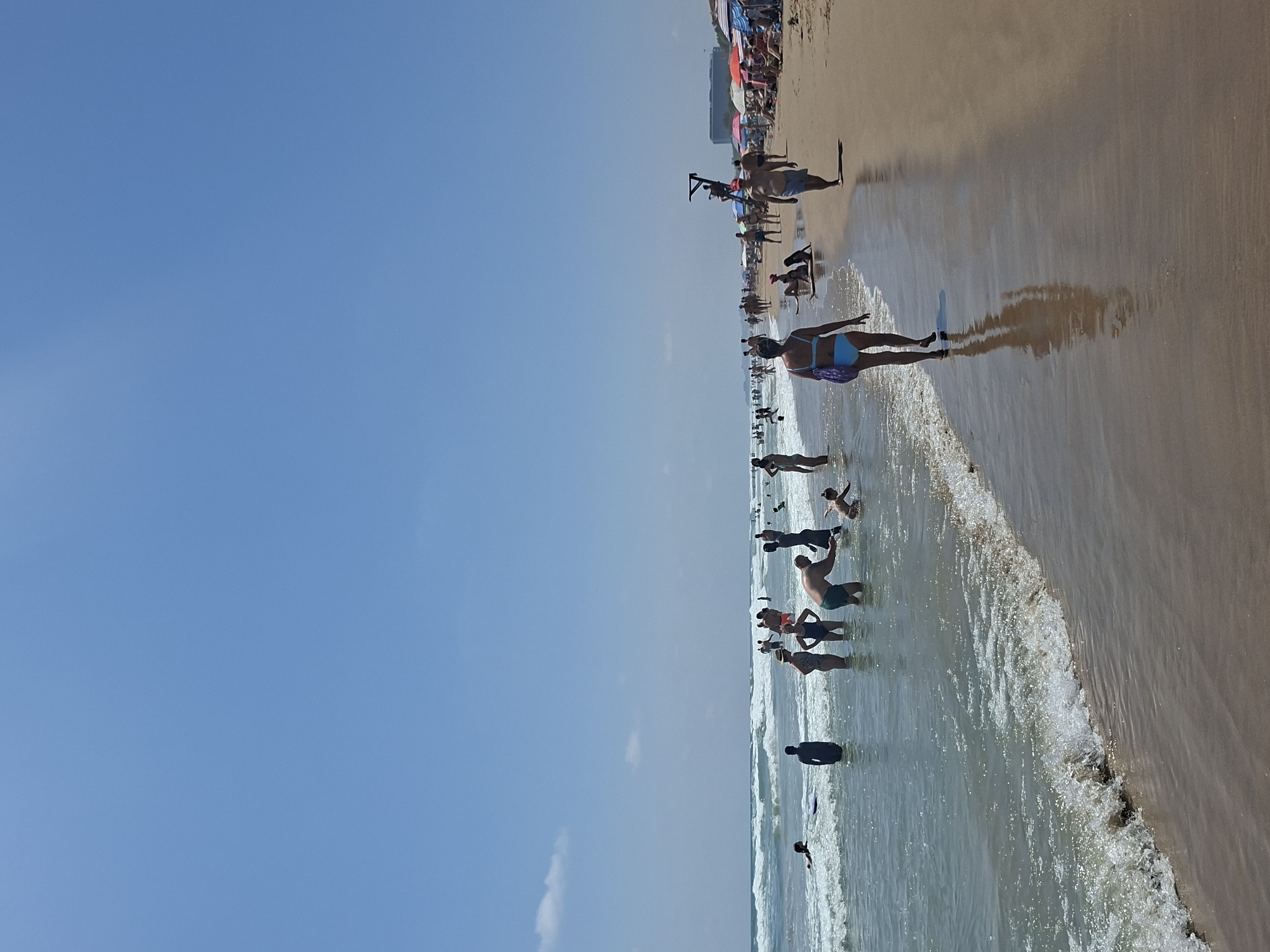 This image depicts a sunny day at a crowded beach in Guardamar del Segura, Spain. The foreground shows the wet sand of the shoreline with gentle waves lapping at the beach. In the mid-ground, several people are in the shallow water, some wading and others swimming. A few individuals are visible further out in the sea. On the sand, more people are spread out, some are walking along the shoreline, others are sunbathing under umbrellas, and a lifeguard is visible on a tall stand. The background reveals a hazy horizon with buildings and a clear blue sky. The overall scene conveys a typical summer day at a popular beach destination.