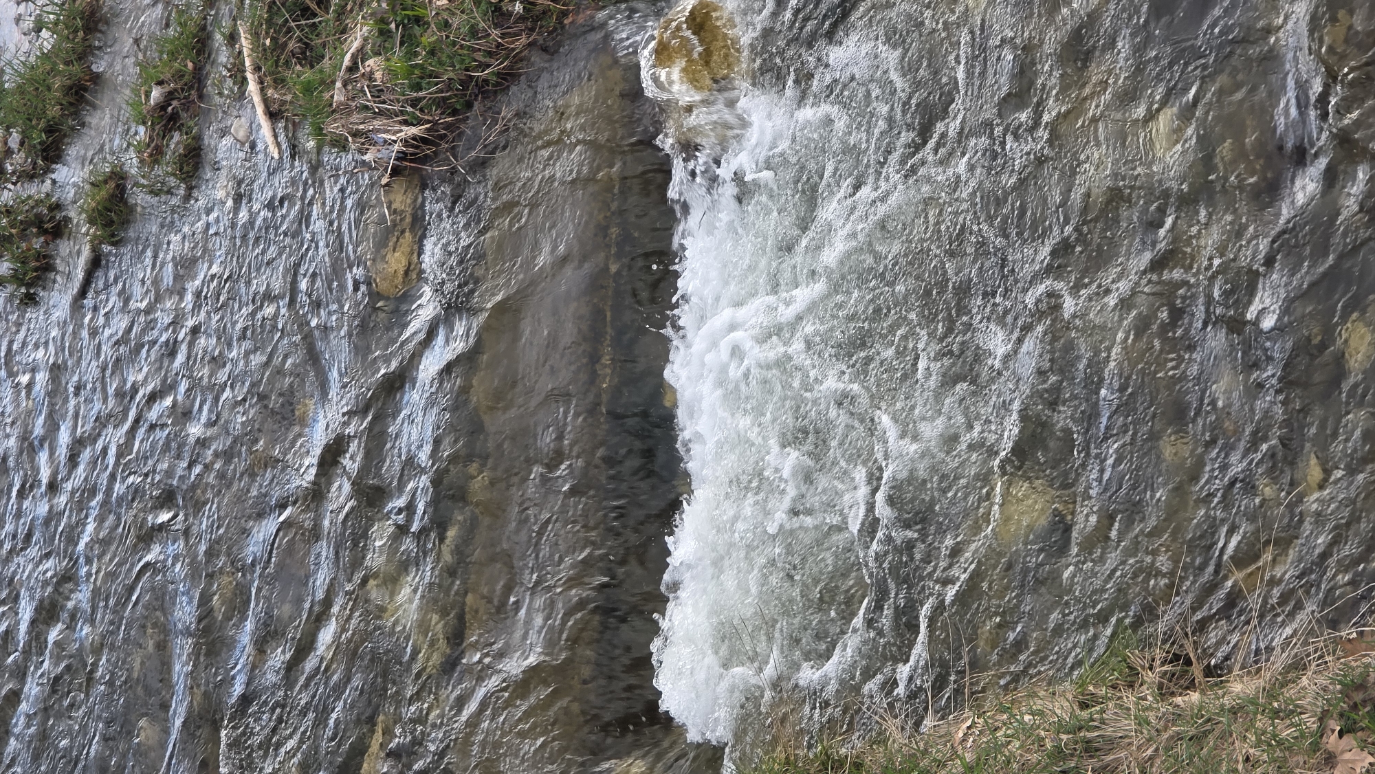 A stream of water flows downward over a rocky surface. The water is turbulent and frothy on the right side, while the left side is smoother. Moss and small plants are visible along the edges of the rocks, and there is vegetation at the top and bottom of the frame. The image could be of a waterfall or a section of a river with a drop.
The location is Blagoevgrad, Bulgaria.