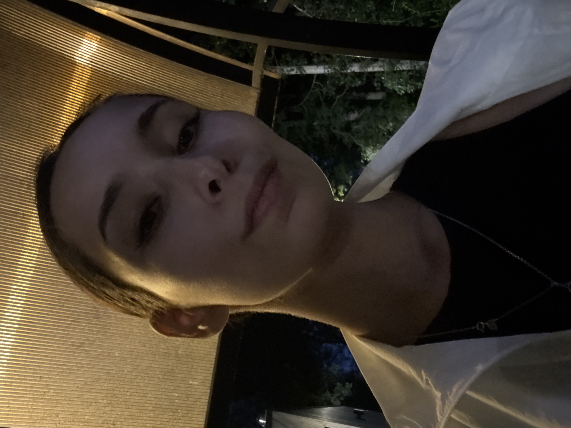 A close-up, low-angle selfie shows a young woman with her hair pulled back and wearing a black shirt and a white jacket. She is indoors, possibly on a patio or under a covered structure, with a slatted ceiling illuminated from above. The background is dark, with trees and foliage visible. The lighting suggests it is evening or nighttime. The woman is looking towards the camera with a neutral expression. The scene is located at Doma 1096 Km, Russia.