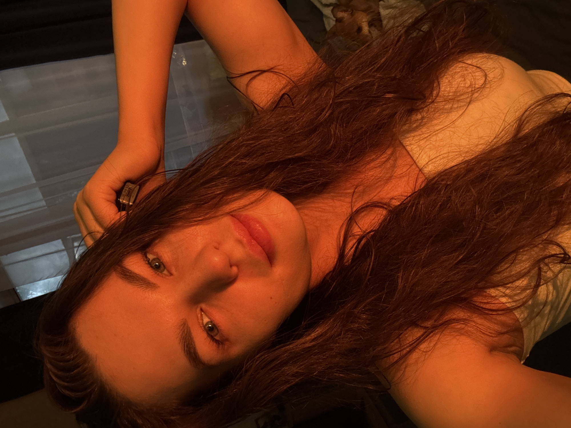 An adult female with long, dark, wavy hair is captured in a close-up, selfie-style shot. She is positioned centrally in the frame, with her head tilted and her left arm raised, hand resting on her head. She wears a strapless top. The lighting is warm and orange-toned, casting a dramatic effect on her skin and hair, and appears to be artificial. The background is indistinct, but suggests an indoor environment with a window covered by curtains on the left and a dark area on the right. A small, light-colored object, possibly a pet, is visible in the lower right portion of the background. The scene is devoid of any identifiable Chaikovsky, Russia landmarks.