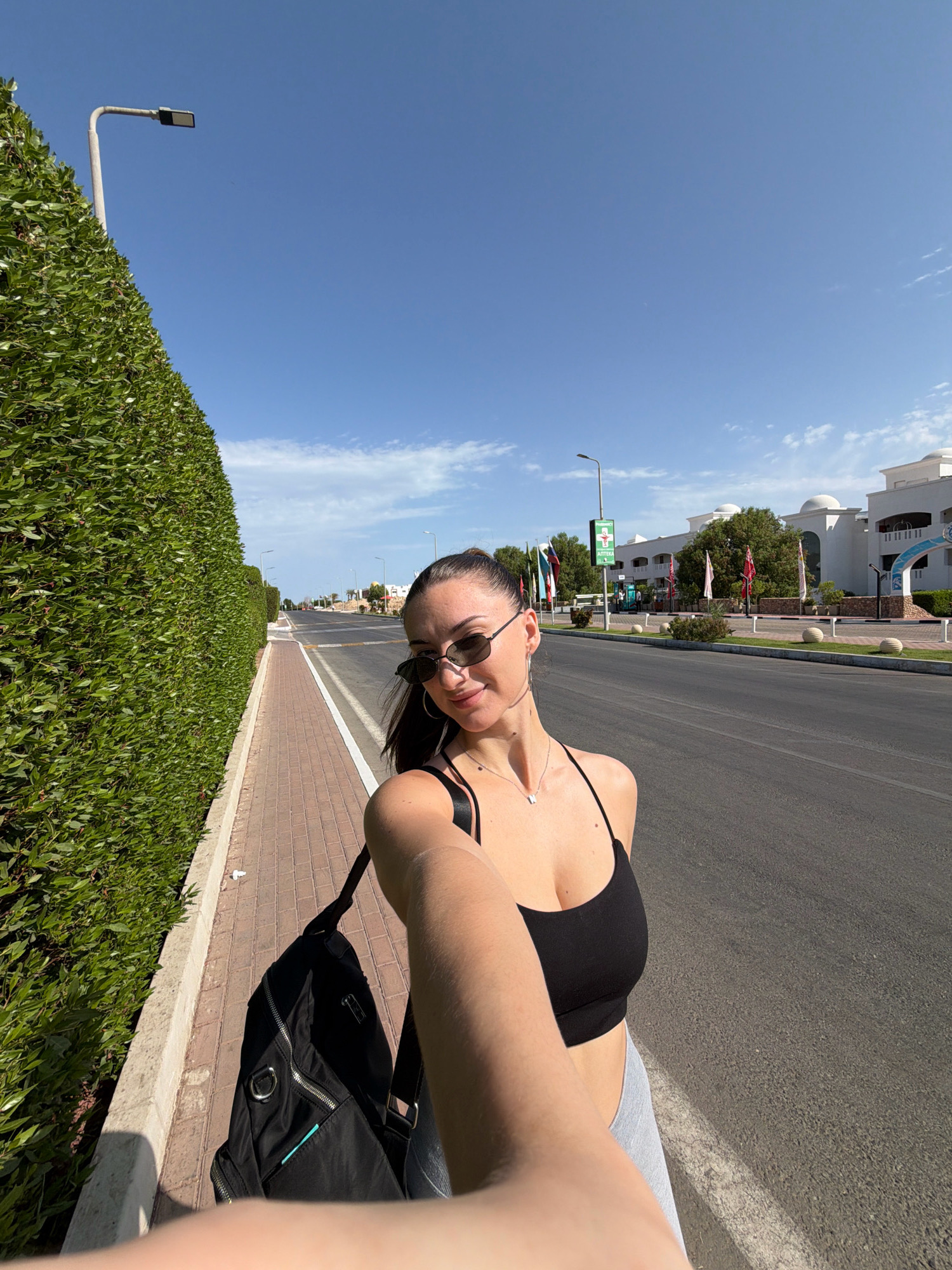 A woman is taking a selfie outdoors on a sunny day. She is wearing a black sports bra and grey leggings, with a black backpack slung over her shoulder. The woman is smiling and wearing sunglasses. She is standing on a paved sidewalk next to a tall, manicured green hedge.
Behind her, a road stretches into the distance, lined with streetlights and more hedges. On the other side of the road, white buildings with domed roofs are visible, suggesting a resort or hotel area. Flags are flying in front of the buildings. A green sign with text, possibly indicating a pharmacy or services, is mounted on a pole. The sky is clear and blue with a few wispy clouds. The scene appears to be in a warm climate, likely during the day. The overall impression is of a casual outing in a vacation destination.