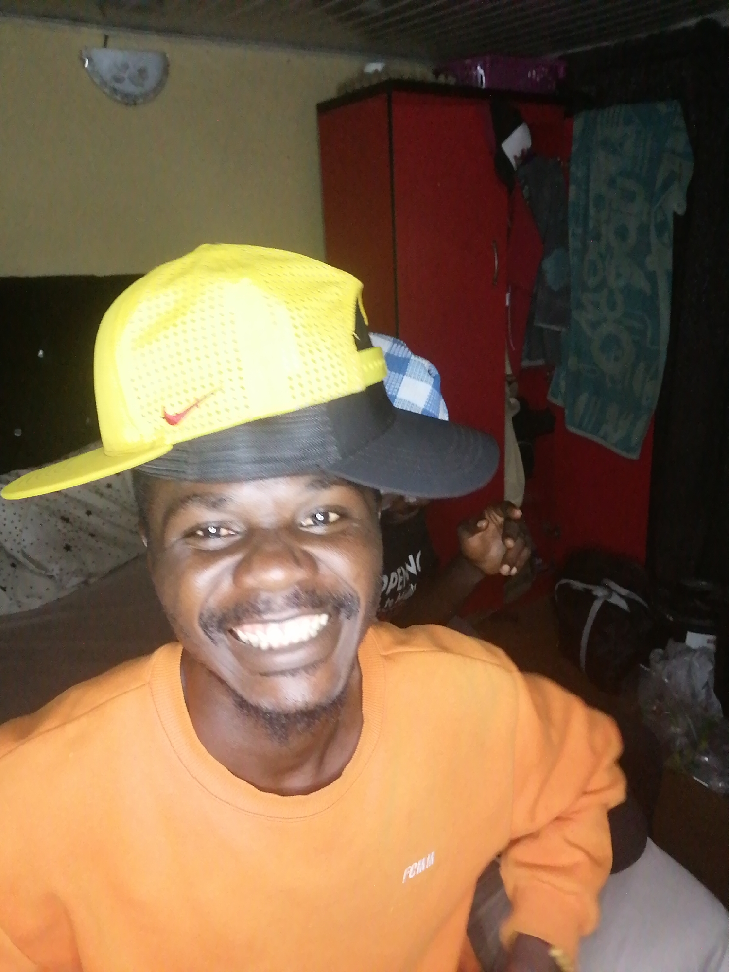 A person is posing for a selfie in an indoor location, possibly a bedroom. The individual is wearing a bright orange sweater and two baseball caps stacked on top of each other: a yellow cap with a red Nike swoosh on top, and a black cap underneath. The person has dark skin, a mustache, and is smiling broadly, revealing their teeth. Behind the person, there is a red cabinet, a patterned towel hanging, and a duffel bag. The setting appears to be a modest living space, with a bed visible in the foreground. There is a light fixture on the ceiling. The location is identified only as an unknown area within the city of Unknown.