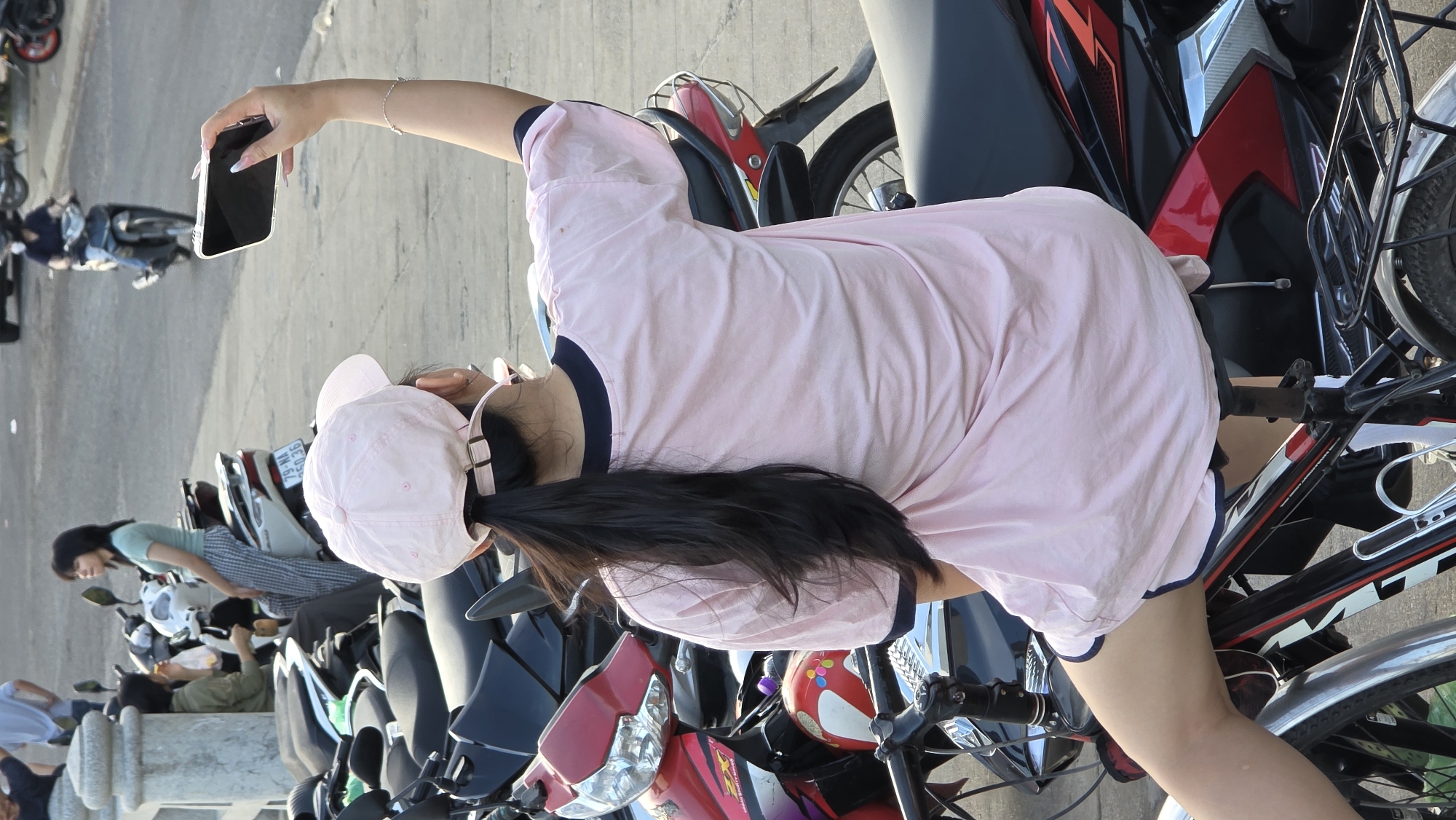 The image depicts a bright, daytime street scene in Bac Nha Trang Ward, Vietnam, centered on a young woman taking a photo with her smartphone. The image, originally rotated, is described as if correctly oriented.
In the foreground, a young woman is seen from behind and slightly to her left. She wears a light pink t-shirt with navy blue trim, matching light pink shorts, and a light pink baseball cap. Her long, dark hair flows down her back. She holds a black smartphone with a clear case in her raised right hand, seemingly capturing an image or video. Her fingernails are painted a light color, possibly with French tips. She appears to be standing or sitting on a dark-colored motorcycle with red accents, whose seat, handlebars, and frame are partially visible. To her right, parts of a silver mudguard and bicycle-like components are visible.
In the mid-ground, a row of motorcycles and scooters is parked along what appears to be a paved street or sidewalk. Another woman, wearing a light blue top and patterned pants, is visible to the left, near or on one of these parked scooters. Further back, another person in a light blue shirt is partially seen. In the distant background, a person on a dark motorcycle rides away.
The setting is an urban or semi-urban environment, characterized by the numerous parked and moving motorcycles, typical of Vietnam. The bright, clear lighting suggests it is sunny daytime. The overall activity appears casual, with people going about their daily routines.
Visible text includes "79-NA 5035" on a license plate of a parked scooter in the mid-ground, and "RS" along with other stylized lettering on the red motorcycle in the foreground, below the main subject's arm. The scene conveys a relaxed atmosphere without any strong emotional cues from the visible subjects.