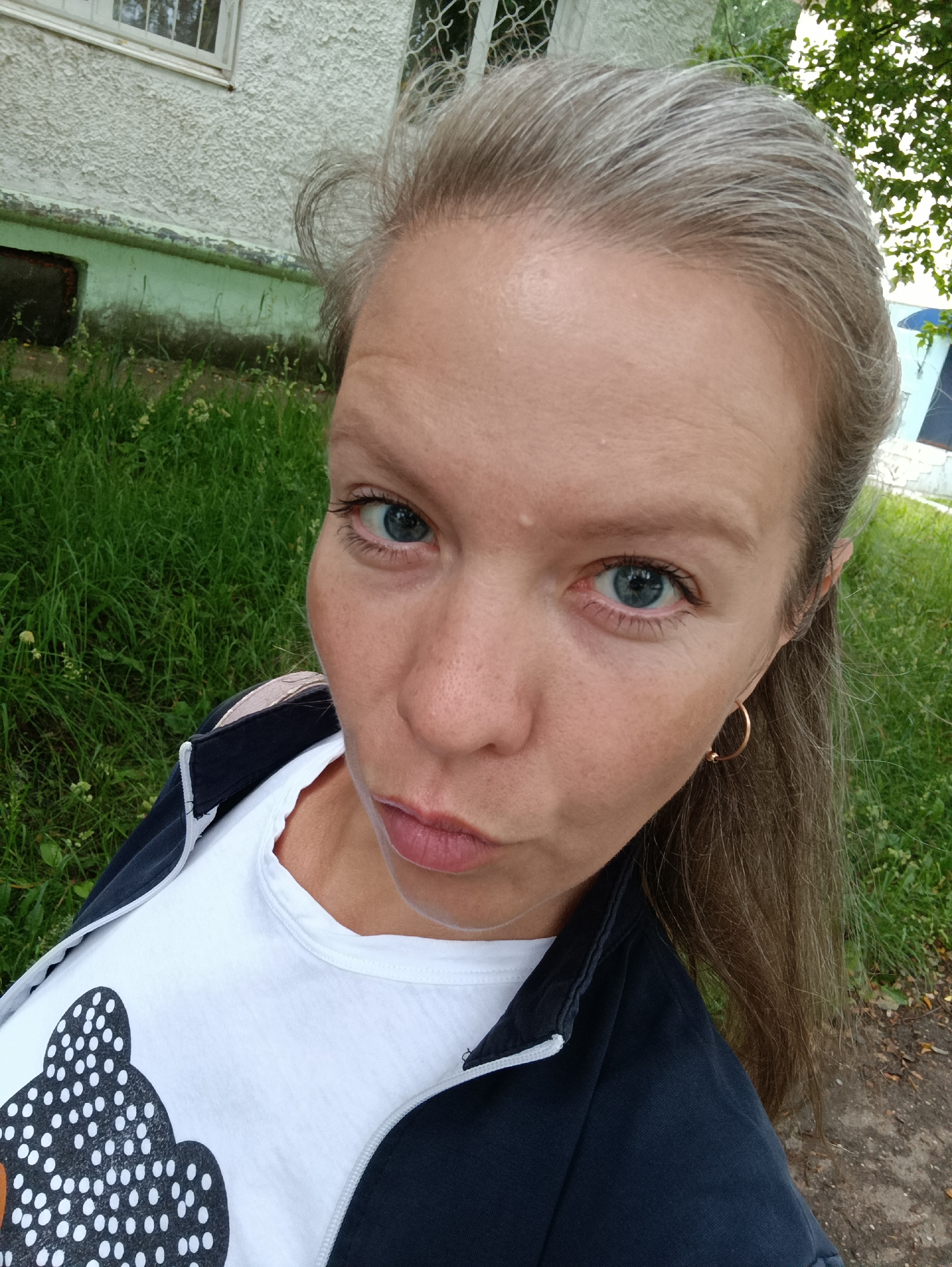 In this close-up, high-angle selfie taken outdoors, a woman with light brown hair and blue eyes is featured. She is wearing a white t-shirt with a black and white dotted design of what appears to be a bear's face, and a dark blue jacket with white trim. She is looking directly at the camera with her lips pursed. The background shows a green grassy area with some trees and a building with a textured white facade and a window. The scene is consistent with a residential area in Chaikovsky, Russia, possibly near a school or apartment complex given the building's appearance and the presence of trees and grass.