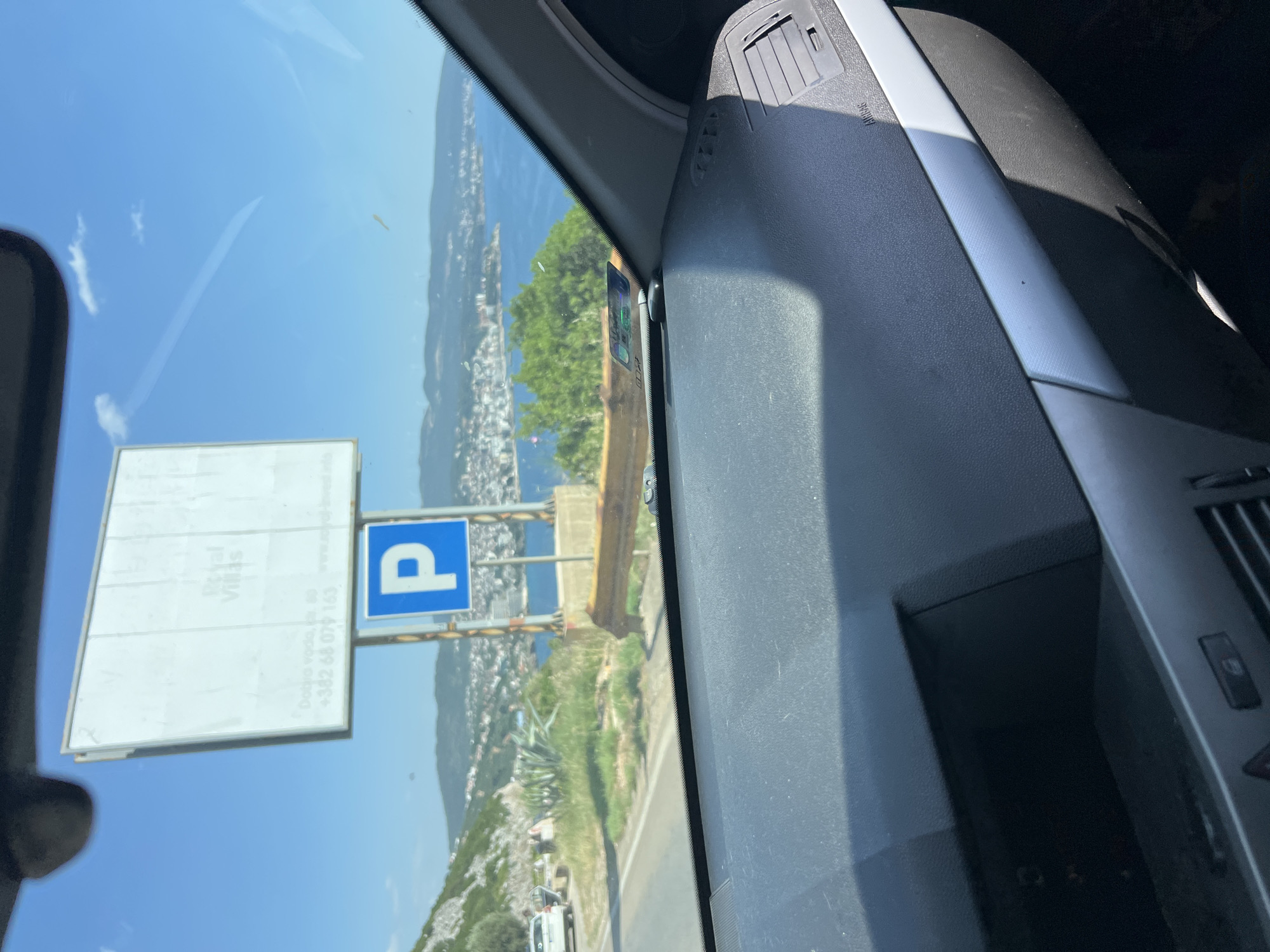 This is a photo taken from inside a vehicle, looking out through the windshield. The scene is a coastal view with a town built along a hillside overlooking the sea. The sky is bright blue with a few wispy clouds, suggesting it is a sunny day, likely in the late morning or afternoon.
In the foreground, the interior of the car is visible, including a dashboard with an air vent and the "AIRBAG" indicator. A large white billboard with some text is positioned to the left, partially obscuring the view of the town. To the right of the billboard is a blue and white sign indicating parking with a white "P" on a blue background.
The town is a cluster of buildings extending down to the water, with green trees interspersed among them. The sea is a deep blue. The overall impression is one of a scenic, perhaps tourist-oriented location.
There is visible text on the billboard and the parking sign. The billboard appears to have text in what might be a foreign language, and below it, there is text that reads: "Dobro voda, ul. 60" and a phone number "+382 68 074 163". The parking sign has a white "P" on a blue square.
The photo is taken at an angle, indicating the vehicle is likely in motion, possibly driving along a road that overlooks the town and the sea. The sunlight suggests a warm, pleasant day. There are no people prominently visible, but the setting appears to be a populated area.