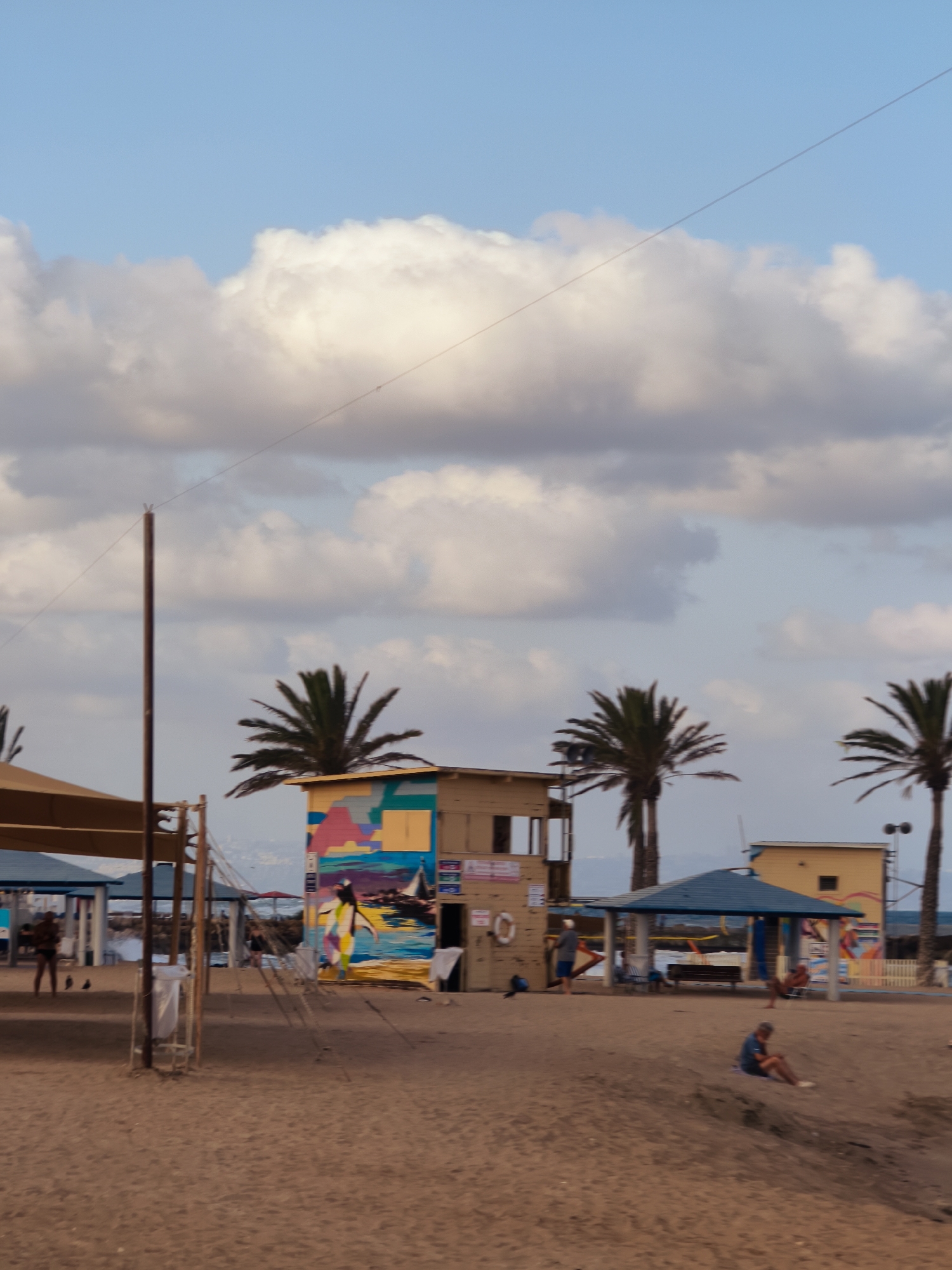 The image depicts a beach scene in Nahariyya, Israel. In the foreground, a sandy beach is visible, with some people sitting or walking. A person is seated on a small sandy mound on the right side of the frame. Further back, a lifeguard station with colorful graffiti adorns its walls stands prominently. Palm trees are scattered across the scene. A yellow building with a blue roof is situated behind the lifeguard station. In the distance, the sea is visible, with a clear sky and scattered clouds above. There are several structures that appear to be beach amenities, such as shaded seating areas and other small buildings. The overall atmosphere suggests a relaxed day at the beach.