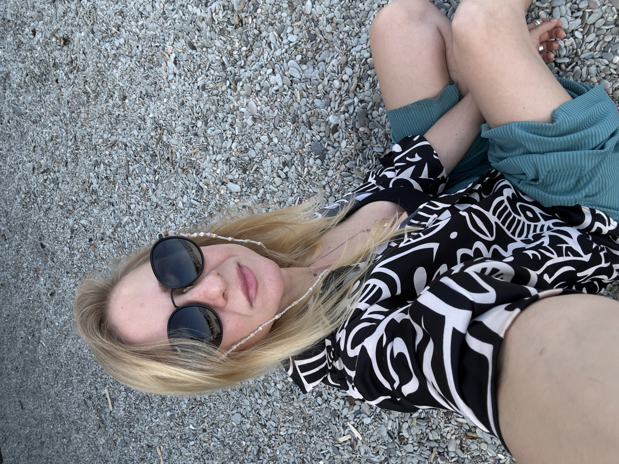 A woman with blonde hair, wearing black sunglasses and a black-and-white patterned top, is lying on a pebble beach. She is also wearing teal clothing. The image appears to be taken from an overhead angle. The location is Unknown, Unknown.