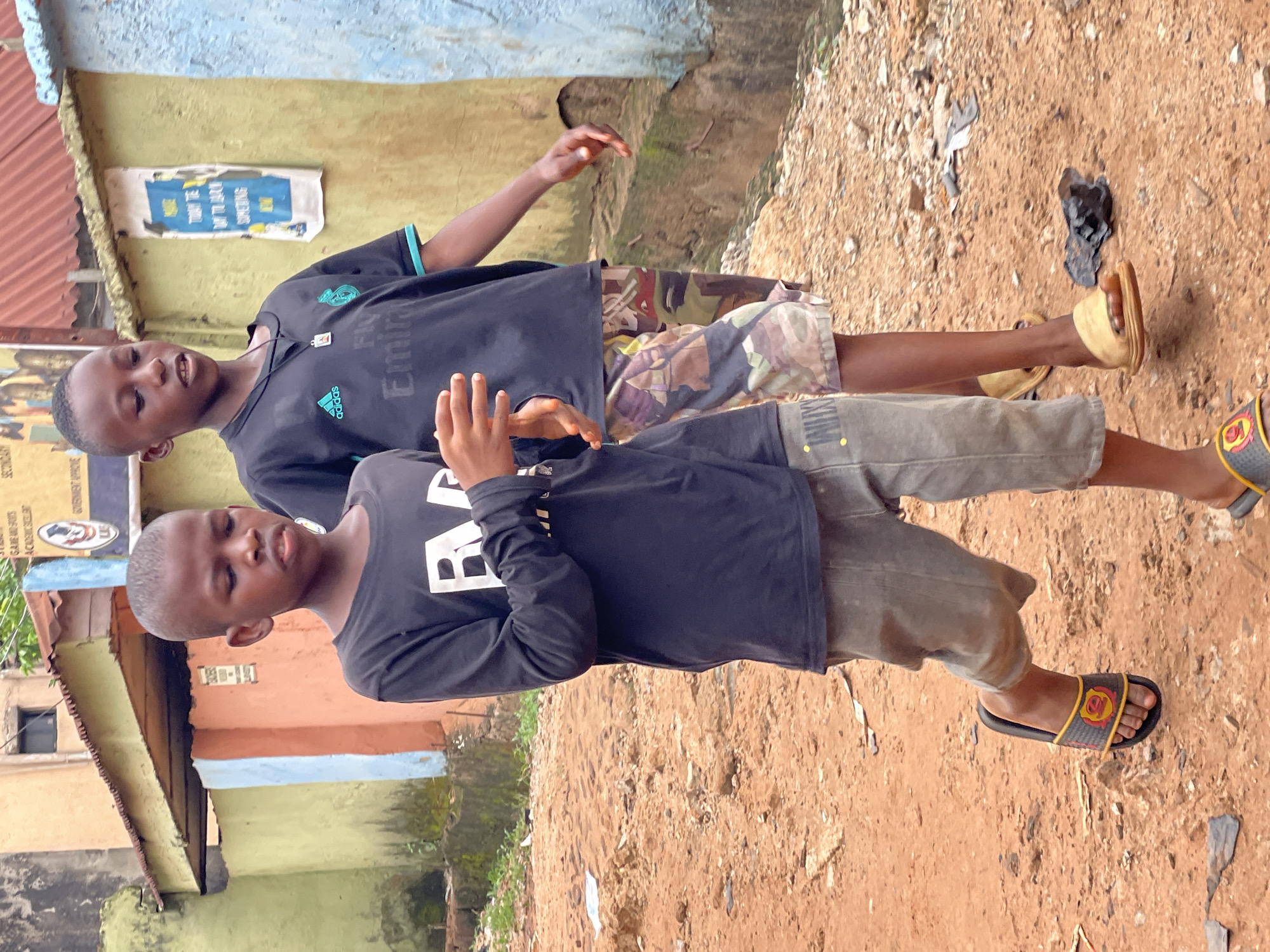 In Nkpor, Nigeria, two young boys are standing outside near a building. The boy in the foreground wears a black long-sleeved shirt, grey pants, and sandals. He has his hands raised. The boy behind him wears a black short-sleeved shirt with an Adidas logo, camouflage shorts, and yellow sandals. The ground is dirt and gravel. There are buildings in the background with signs and faded paint.