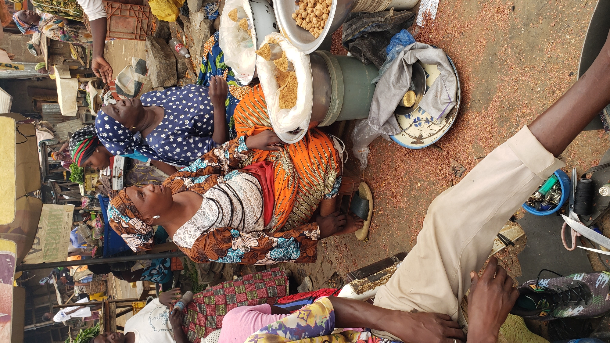 The image shows a market scene in Narayi, Nigeria. Two women are seated on the ground next to a green metal barrel. On top of the barrel is a scale with bowls and bags of what appear to be grains and beans. A third woman is seated behind the first two and a man in a white shirt is visible on the left. A man in a tan shirt is sitting on the right. Debris is scattered on the ground.