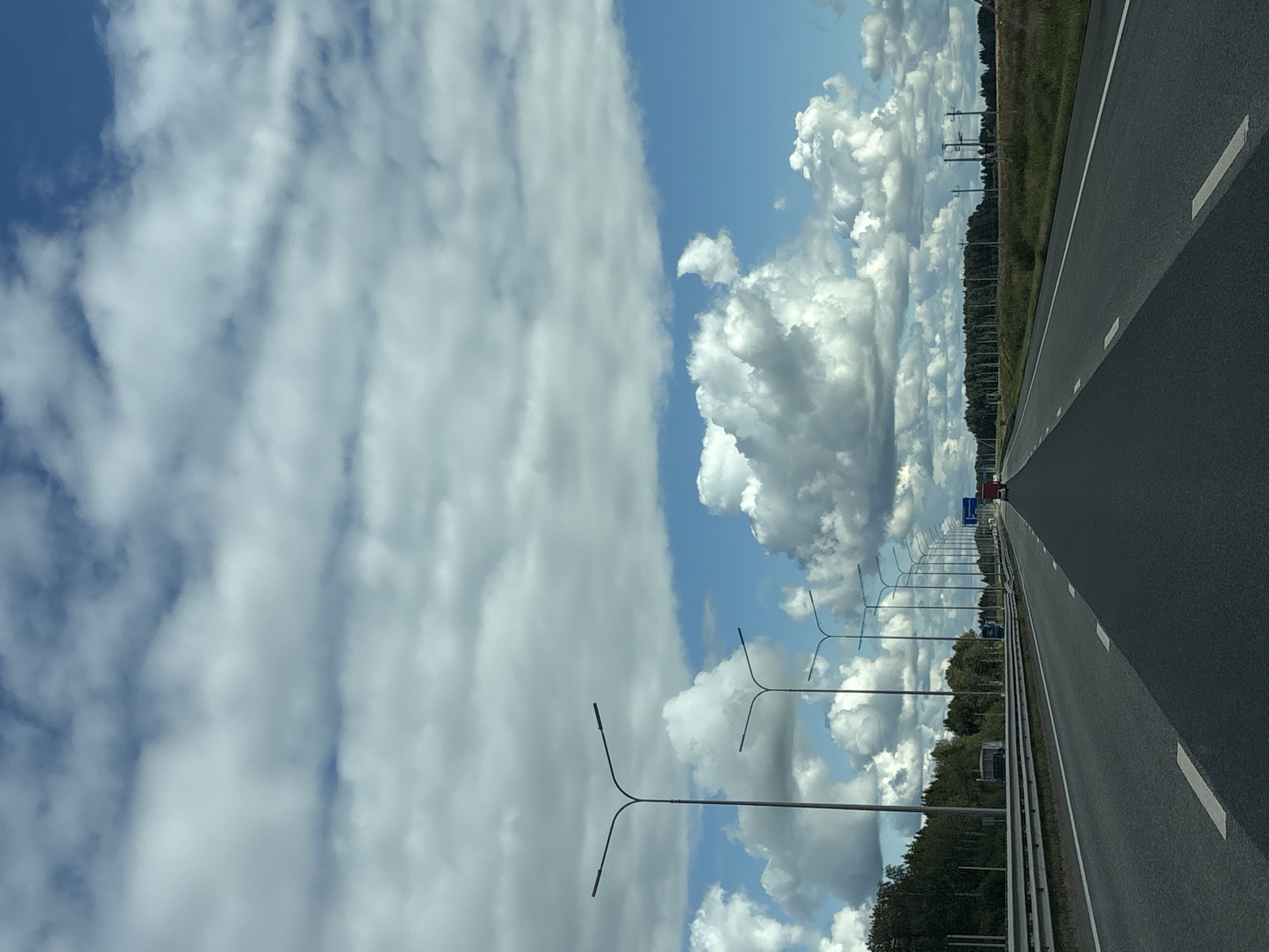 The image depicts a view from a vehicle traveling on a highway. The road has multiple lanes and is paved with asphalt, marked with white lines. On the left side of the road, there is a metal guardrail, followed by a treeline and then fields. Several tall streetlights with modern designs are visible along the highway. In the distance, there are more trees and what appear to be utility poles. The sky is partly cloudy with large, white cumulus clouds against a bright blue sky. A red truck is visible in the far distance on the right lane of the highway. The scene suggests a rural or suburban highway environment. The metadata indicates the location as Yanychi, Russia.
