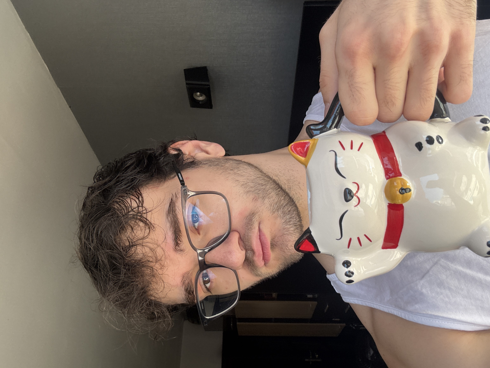 A man with dark hair and glasses is holding a white ceramic maneki-neko mug. He is wearing a white t-shirt. A black speaker is visible in the background. The photo appears to be taken indoors. The location in Blagoevgrad, Bulgaria cannot be determined from the image.