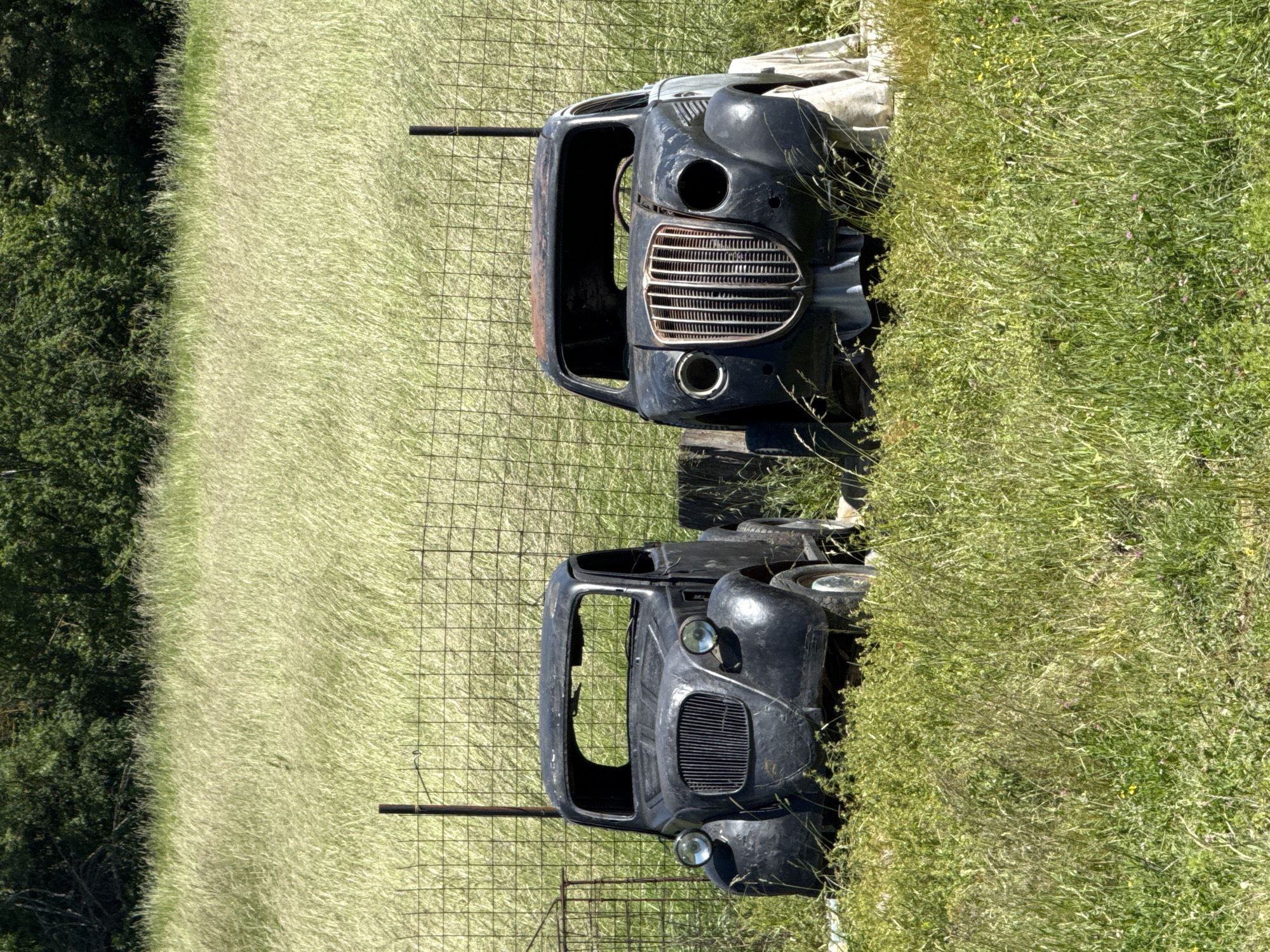 The image shows two derelict car bodies in a grassy field. The vehicles are dark-colored and appear to be vintage models, possibly from the mid-20th century. Both cars lack windows, doors, and likely engines. One car still has wheels visible. A wire fence runs through the field, separating the cars from the grassy area on the other side. The scene conveys an atmosphere of abandonment and decay, with the car bodies being reclaimed by nature. The location is Omurtag, Bulgaria, and is characterized by a grassy field, possibly on the outskirts of the city.