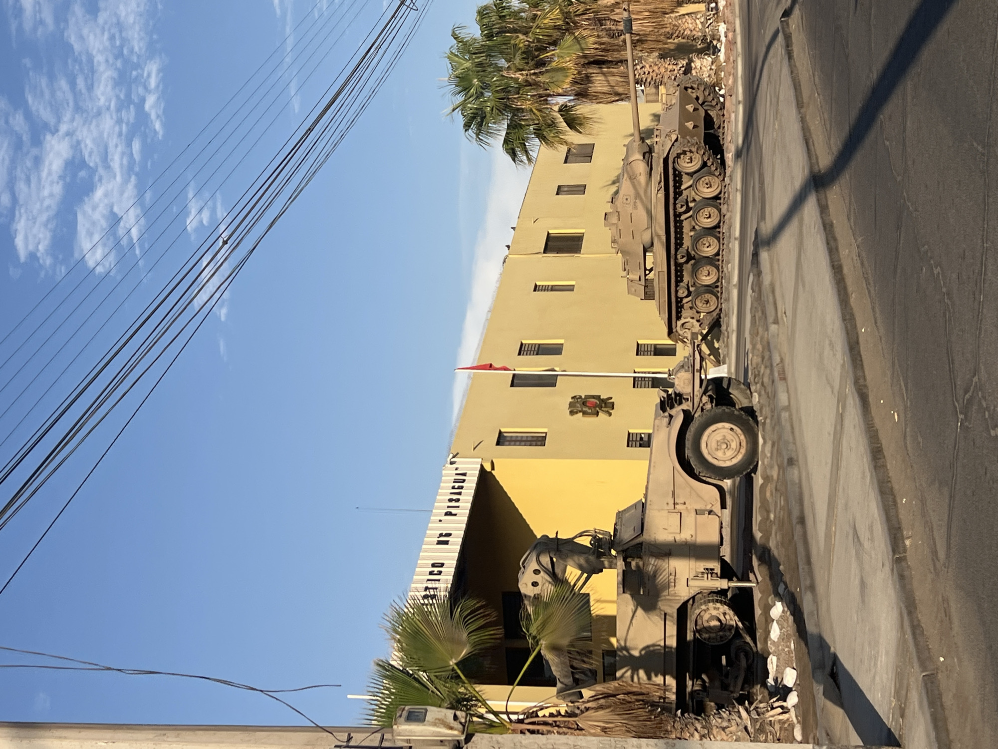 The image shows two military vehicles, a tank and an armored personnel carrier, parked on a street in Arica, Chile. The vehicles are located near a yellow building with the inscription "Atletico N° PISAGUA." A red flag is visible atop the building. Palm trees are situated nearby. Overhead, power lines are strung across the blue sky, dotted with clouds. The street appears to be paved and shows signs of wear and tear.