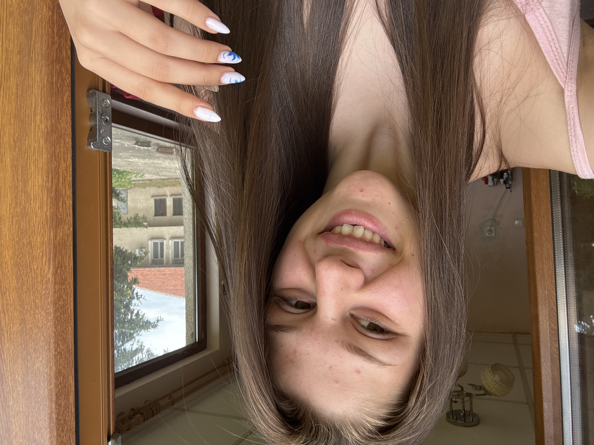 A young woman with long, brown hair and light skin smiles at the camera. She is wearing a pink tank top. Her nails are painted white with blue swirls. She is standing in front of a wooden doorframe and a window that looks out onto a street with a house and trees. A light fixture is visible on the ceiling.