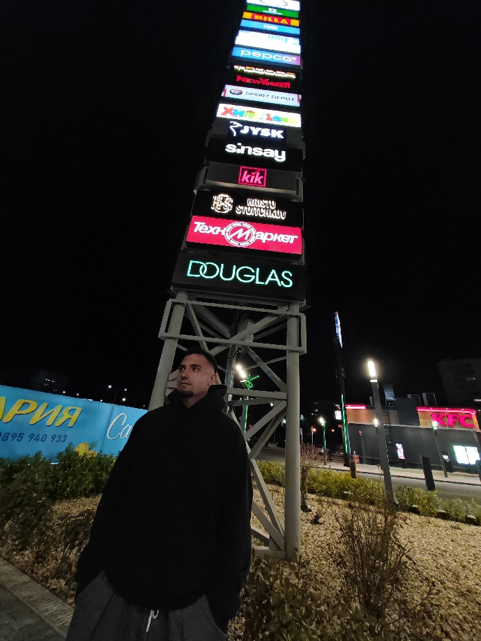 This nighttime image depicts an adult male standing in a retail park in Shumen, Bulgaria. The man, with short dark hair and a trimmed beard, wears a dark oversized hooded sweatshirt and dark sweatpants, looking upwards and to the right of the frame. He is positioned in front of a tall, vertical, multi-tiered signpost constructed from grey metal scaffolding. This signpost features numerous horizontally stacked, illuminated brand logos, including (from top, partially visible) BILLA, Pepco, New Yorker, Sport Depot, Hippoland (ХИПОЛЕНД), JYSK, Sinsay, KiK, Hristo Stoichkov (ХРИСТО СТОИЧКОВ), Technomarket (Техномаркет), and Douglas.
The ground around the man and the signpost is covered with light-colored gravel or small stones and sparse, low-lying vegetation. To the immediate left of the man, a large blue sign or billboard with white Cyrillic text "АРИЯ" and a phone number "0895 940 933" is partially visible. In the background to the right, beyond the signpost structure, a brightly lit KFC restaurant is visible, along with other commercial buildings and modern vertical streetlights. The scene is illuminated by artificial light sources from the signs, buildings, and streetlights against a dark night sky, indicative of an active commercial area.