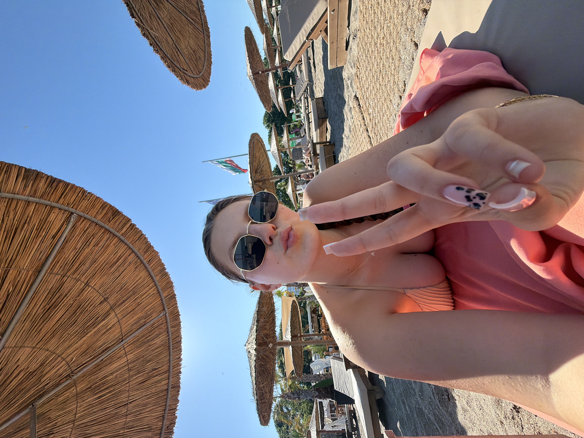 A young woman with blonde hair pulled back in a braid and wearing sunglasses and a beige bikini top is lying on a beach lounger on a sunny day. She is holding up two fingers in a peace sign and puckering her lips for a selfie. The beach is lined with thatched umbrellas and loungers, and the sea is visible in the background. The image appears to have been taken at the northern beach of Burgas, Bulgaria, indicated by the Bulgarian flag flying in the background.