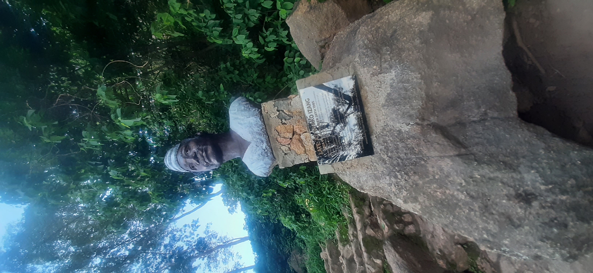The image depicts a bust of a person, possibly a historical figure, positioned outdoors amidst lush greenery. The bust is dark-skinned, wearing a white head covering and a white garment around the neck and shoulders. It is mounted on a rough, textured base that appears to be part of a larger rock formation.
A plaque is attached to the rock base, featuring text and an image. The visible text on the plaque includes "FATE..." and "MARO IZANG" followed by "DECEMBER 26TH 1985". Below this, there is more text, some of which is illegible due to the image quality and angle, but appears to discuss alliances and historical significance. The image on the plaque shows a black and white scene, possibly a historical event or location, with trees and structures.
The surrounding environment is characterized by dense foliage and large rocks, suggesting a natural or park-like setting. The lighting suggests it is daytime, with soft illumination and shadows indicating the sun is likely not directly overhead. The overall mood is contemplative and historical, with the bust and plaque serving as a memorial or marker. The location context of Jos, Nigeria, provides a specific cultural and geographical backdrop for this monument.