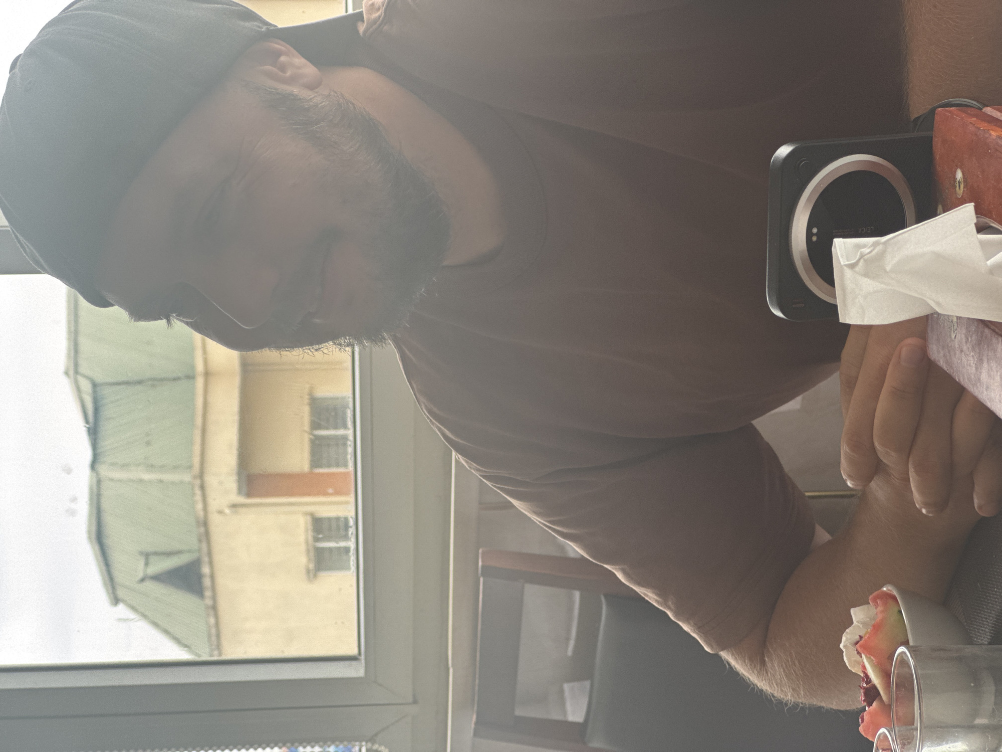 The image shows a medium shot of a man sitting at a table, with his head turned to the side, looking out a window. He is wearing a dark baseball cap and a brown t-shirt. His hands are clasped in front of him, resting on a box of tissues next to a small camera. In the background, a building with a green roof is visible through the window, along with a cloudy sky. To the left of the man, a bowl of fruit salad and a glass of water are visible on the table. The environment appears to be indoors, possibly a restaurant or cafe, given the presence of tables, chairs, and a window with a view of urban architecture. The overall lighting is soft and diffused, with some lens flare visible, suggesting the photo was taken during the daytime. The image is composed in a way that focuses on the man's profile and the objects on the table. The quality of the image is slightly soft, with some blurriness in the background. There is no specific indication of the location being Hà Nội, Vietnam, based solely on the visual content.