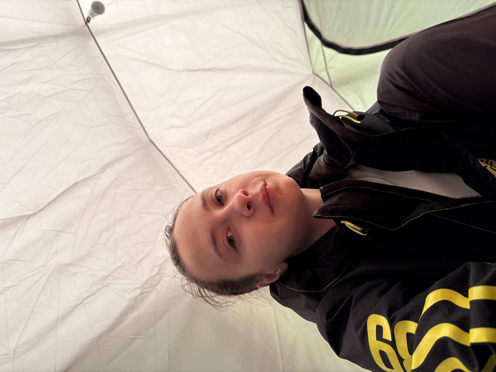 A young woman is looking directly at the camera. She is wearing a dark-colored jacket with yellow accents. The photo appears to be taken inside a tent. The walls of the tent are light-colored and somewhat translucent. There is no discernible evidence to confirm the location in Lesnoye, Russia.