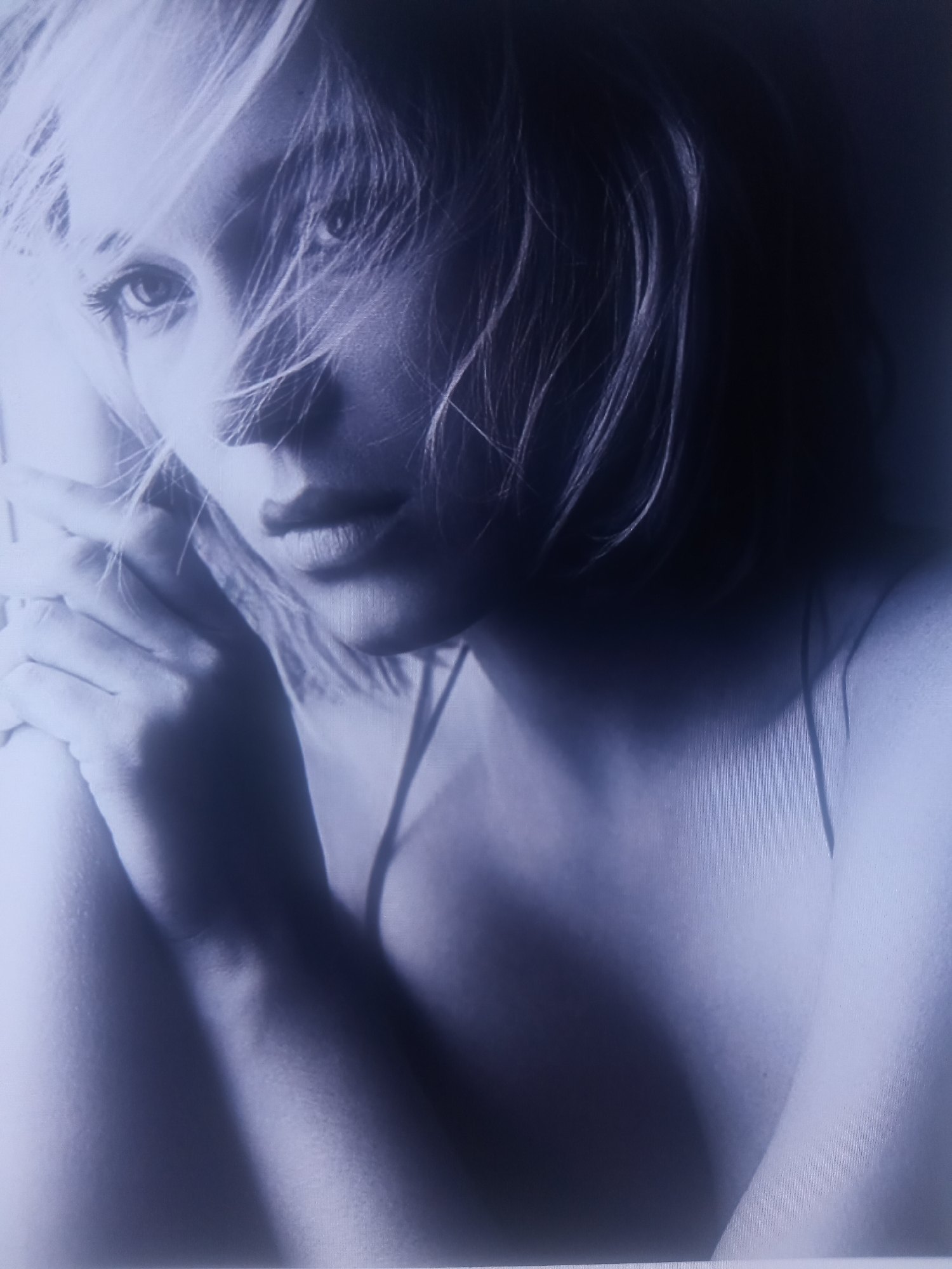 A close-up, grayscale portrait of a woman's face and upper torso is presented. Her blonde hair is swept across her face, partially obscuring her eyes and forehead. Her left hand is raised, with fingers resting near her cheek, and a strand of hair is visible between her fingers. Her lips are slightly parted, and her gaze is directed forward. The lighting creates strong shadows on her face, highlighting the contours of her features. She is wearing a thin-strapped garment, visible on her shoulder and chest. The background is dark and indistinct, with no discernible environmental features.