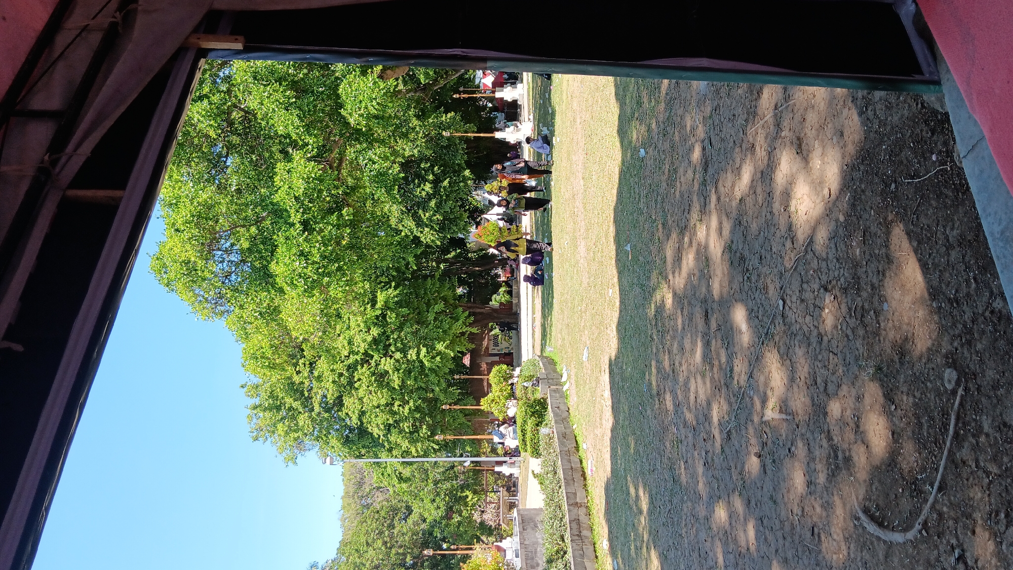This is a vertical, medium-angle shot looking out from a shaded area into a public park on a sunny day in Nganjuk, Indonesia. In the foreground, a dark, textured awning or overhang is visible at the top and right edges of the frame, with a sliver of red fabric at the bottom right. The ground in the immediate foreground is a mixture of dry, cracked earth and sparse grass, with fallen leaves and a few pieces of litter.
Beyond this, a manicured lawn extends into the midground, dotted with several groups of people. The people are primarily observed walking or sitting on the grass. One group of three women in yellow tops and dark headscarves are walking towards the right side of the frame. Another group of two women are walking to the right. Further back, another woman is walking in the same direction. Several other individuals are seated on the grass in small groups.
In the background, a large, mature tree with lush green foliage dominates the scene, casting dappled shadows on the lawn. To the left of this tree, a streetlamp and several ornate lampposts are visible. In the far left background, more trees and buildings are partially obscured. A low stone wall with a manicured garden bed runs across the midground, separating the foreground ground from the lawn. The sky is clear and bright blue. The overall impression is of a casual daytime gathering in a green urban space.