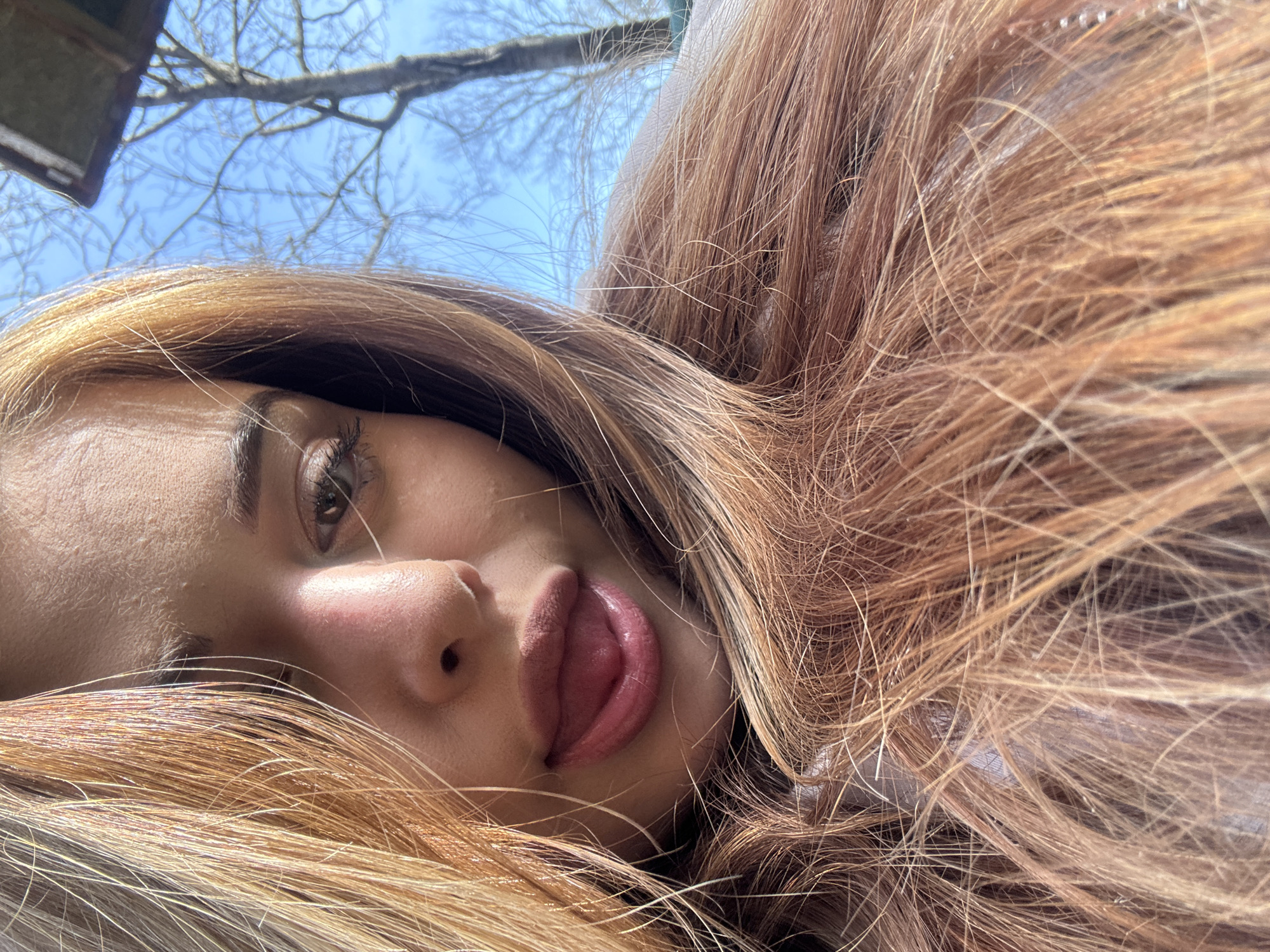 A young woman is lying down, possibly on a blanket or soft surface, with her head at an angle. Her long, wavy hair is spread around her, and her face is tilted towards the camera. She has fair skin, brown eyes, and full lips that are slightly parted, revealing the tip of her tongue. The background features a blue sky and bare tree branches. The location is not discernible from the image, and a determination of whether it is Gorno Trape, Bulgaria, cannot be made.