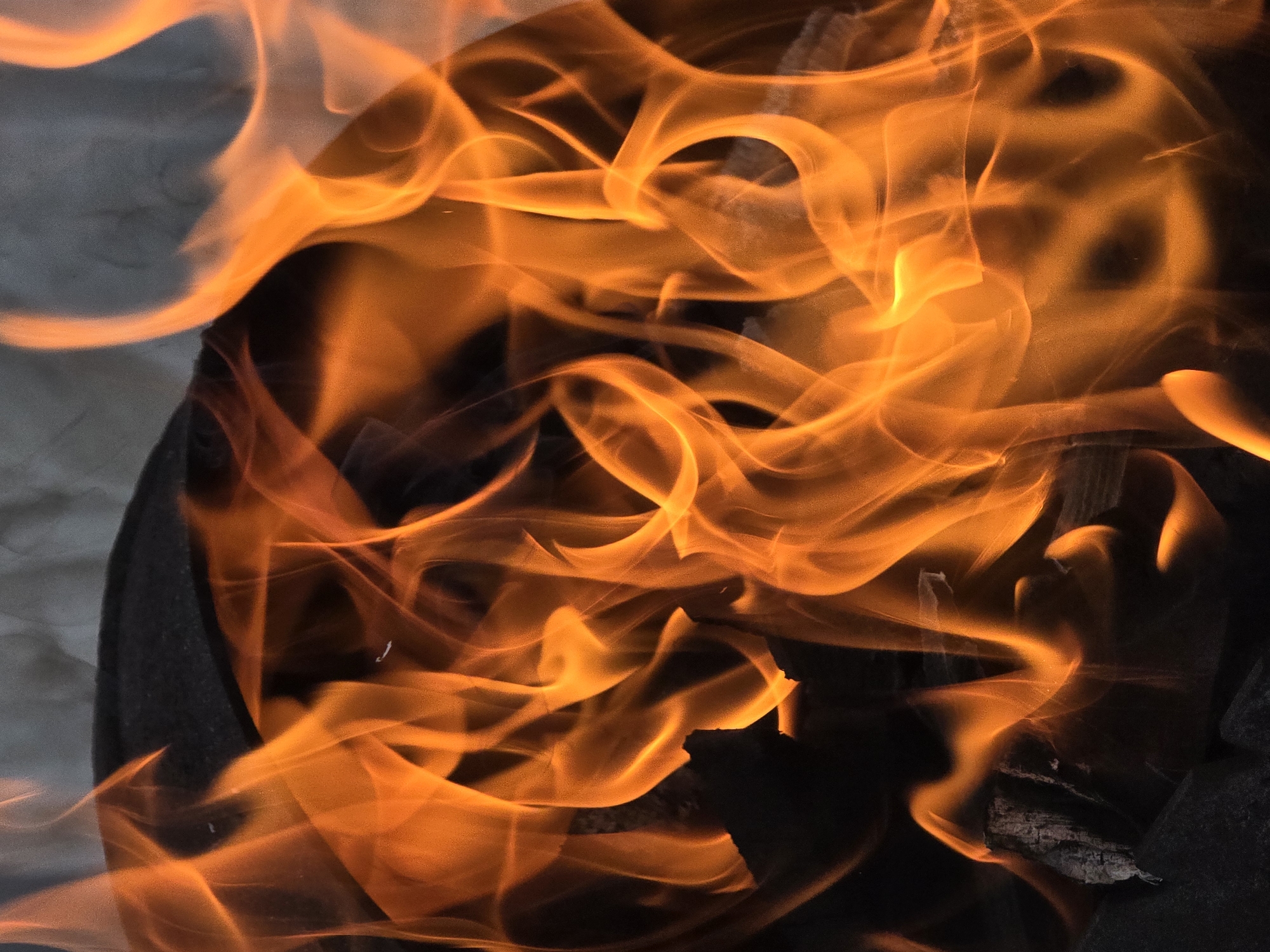 The image shows a close-up view of a fire. Bright orange flames with wisps of yellow and red dance and curl, some reaching upwards and others swirling around. The fire appears to be burning wood, as evidenced by the dark, charred textures visible beneath the flames. The background is a soft, out-of-focus gray, suggesting an outdoor setting or a dimly lit environment. The image is dominated by the vibrant and dynamic movement of the fire, with no discernible people or specific city details.