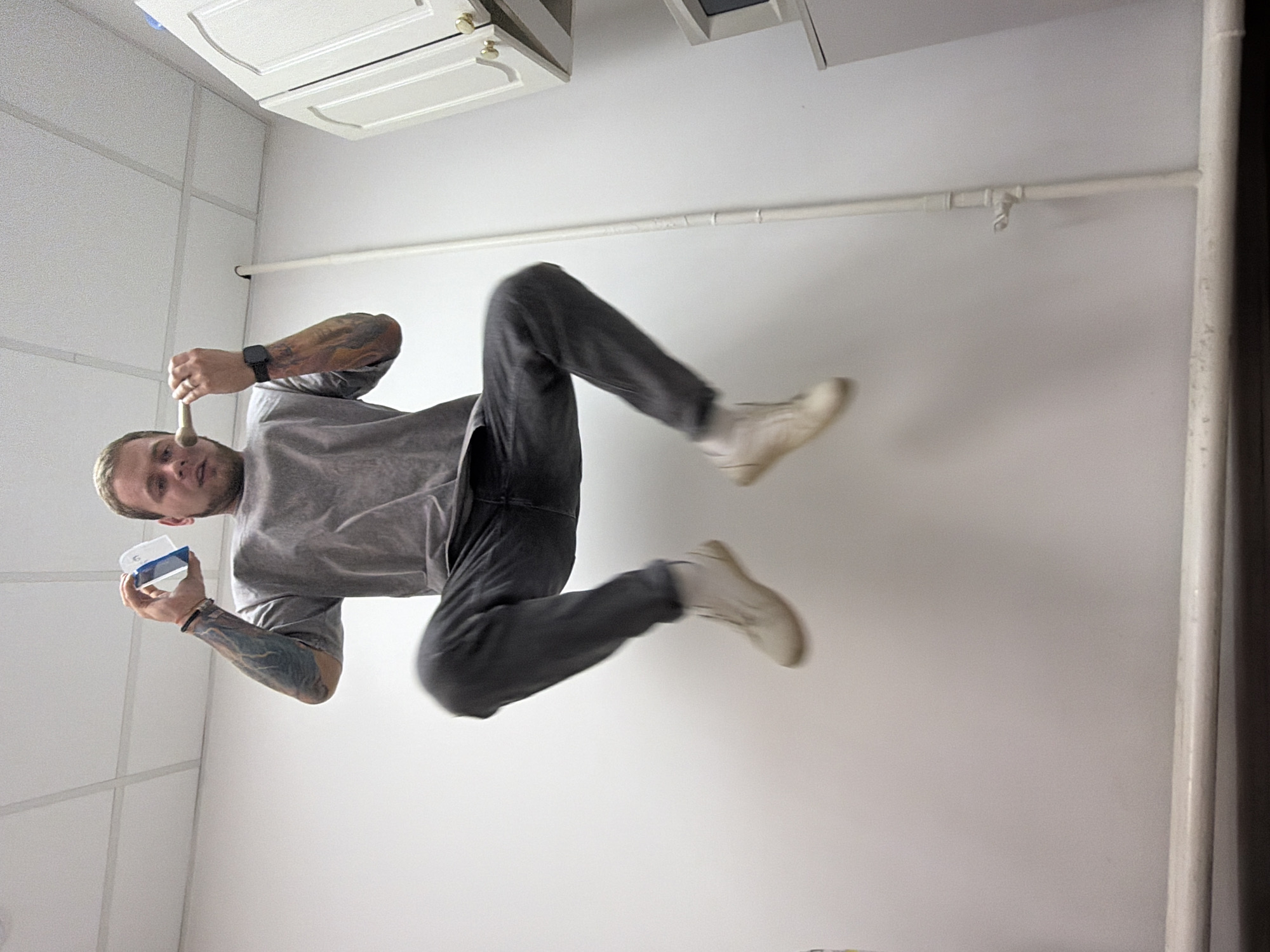 A man with light hair and tattoos on his arms is captured in a motion blur mid-air, as if jumping or falling, inside a room. He is wearing a gray t-shirt, dark jeans, and light-colored boots. In his hands, he holds a small container and a utensil, possibly a spoon or brush. The room has white walls, a white tiled ceiling, and white cabinets. A white pipe runs along the wall. The exact location in the city of Chaikovsky, Russia, is not discernible from the image.