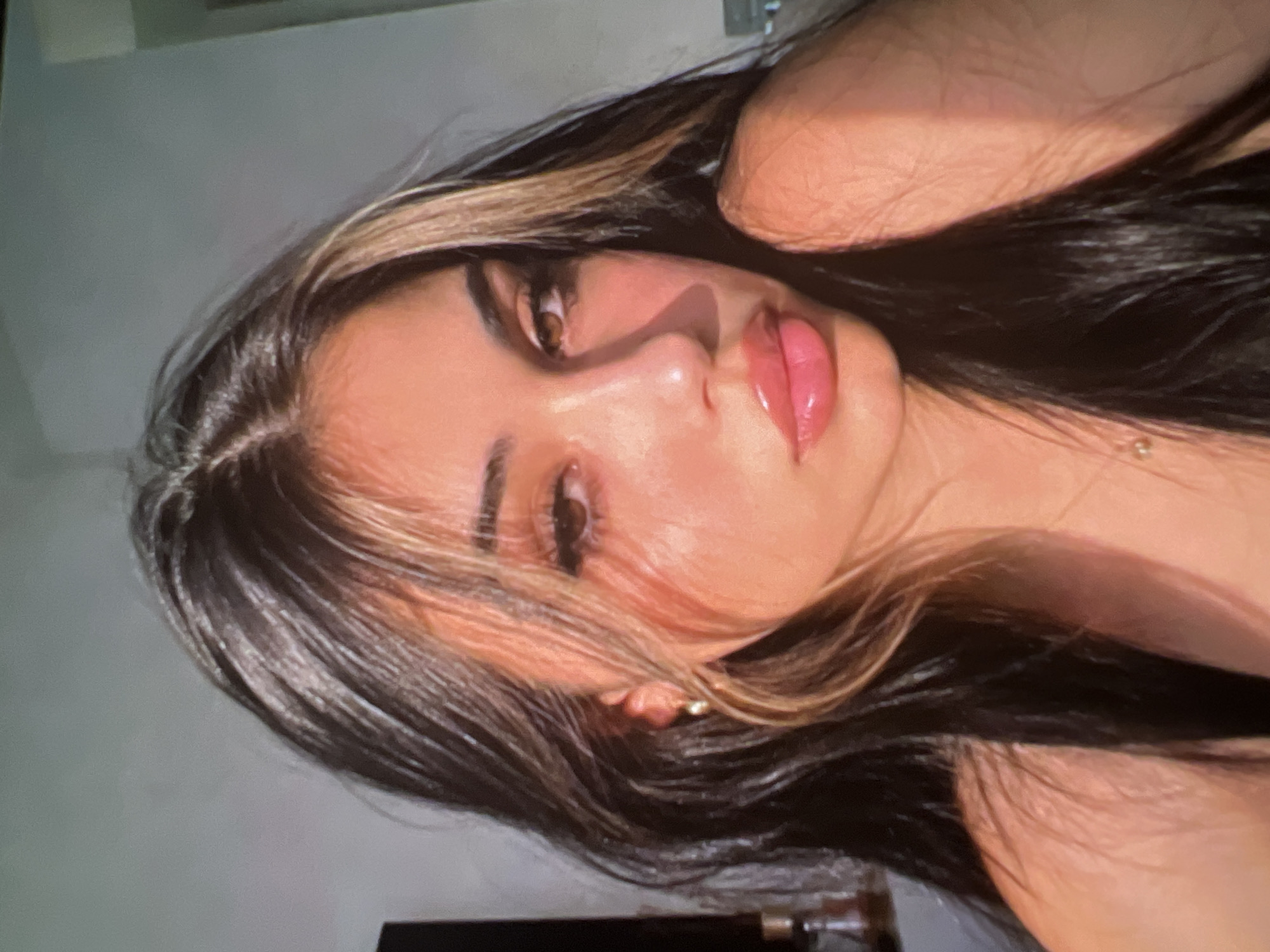 The image is a close-up, head-and-shoulders portrait of a young woman with long, dark hair with blonde highlights. She is looking directly at the camera with her mouth slightly open, revealing plump, glossy lips. Her eyes are brown and her eyebrows are dark and well-defined. She is wearing a gold earring in her left ear and a thin gold chain around her neck. The lighting suggests natural light, possibly from a window, casting shadows on her face and shoulder. The background is a plain, light-colored wall. There are no discernible elements in the image that indicate a specific location in the city of Athens, Greece.
