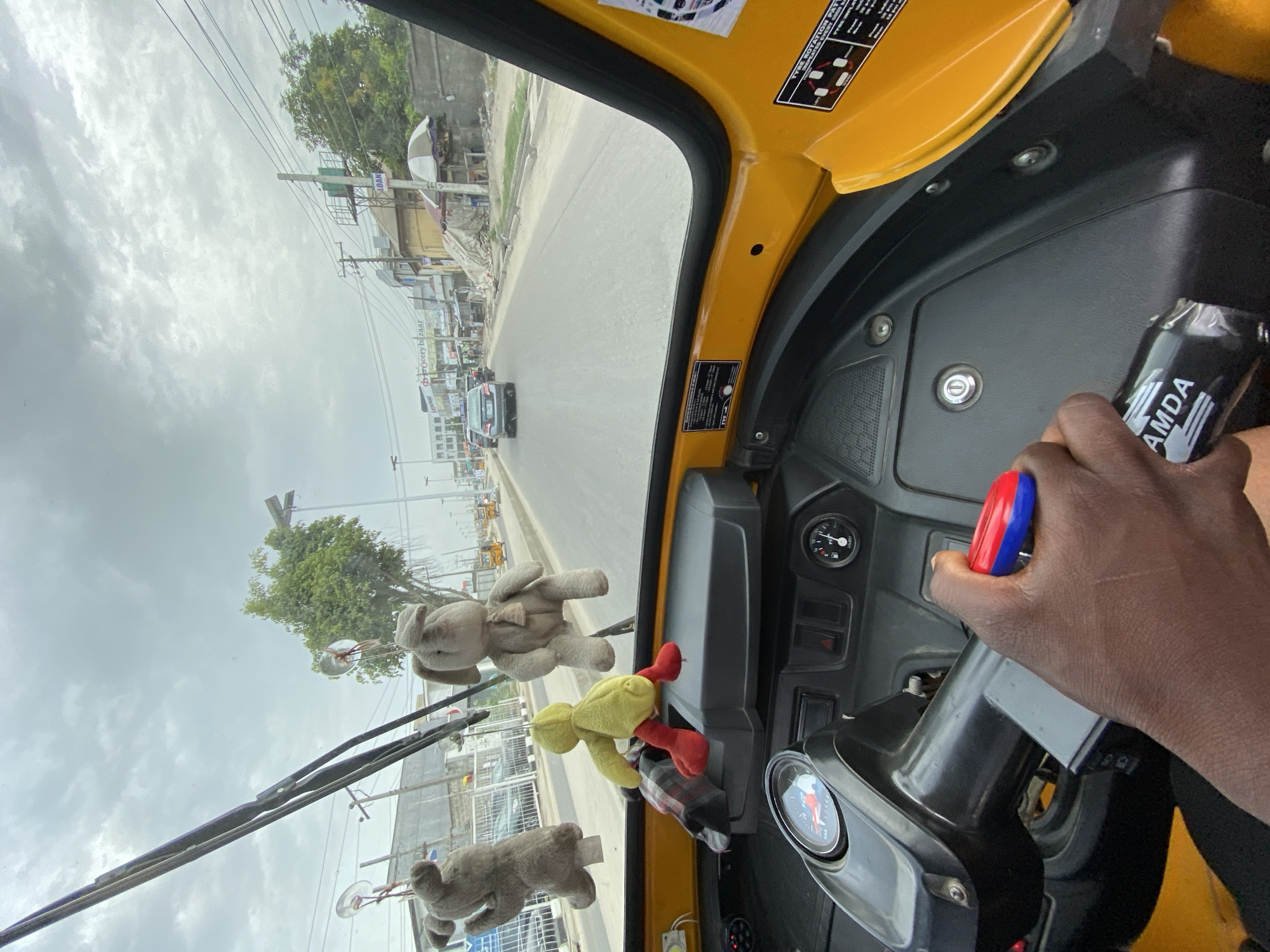 The image shows the interior of a yellow tricycle rickshaw in Ado, Nigeria, facing down a paved road with buildings and power lines visible in the background. The driver's hands are visible, one holding a can of "AMDA" energy drink, and the other on the steering column. Plush toys are hanging from the front of the rickshaw. A car is visible in the distance on the road. The sky is overcast.