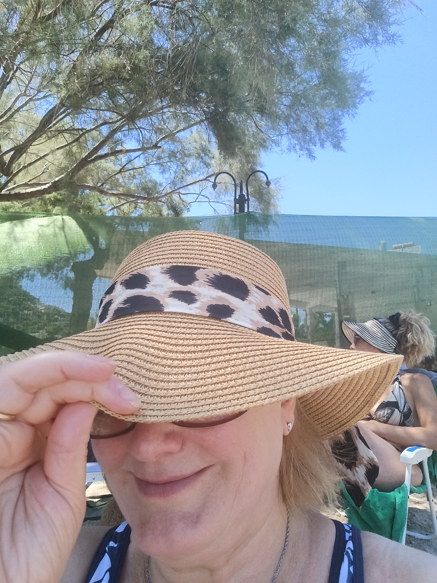 In Agia Marina, Greece, a close-up selfie shows a woman wearing a wide-brimmed straw hat with a leopard print band. She is holding the brim of her hat with her left hand, obscuring her eyes. Her head is tilted slightly upwards, and she is smiling. She is wearing a silver necklace. In the background, partially visible, is another person sitting in a chair, wearing a striped sun visor and a patterned swimsuit. Behind them, a green mesh screen is set up, with trees and a clear blue sky visible above. The setting appears to be a beach or outdoor recreational area.