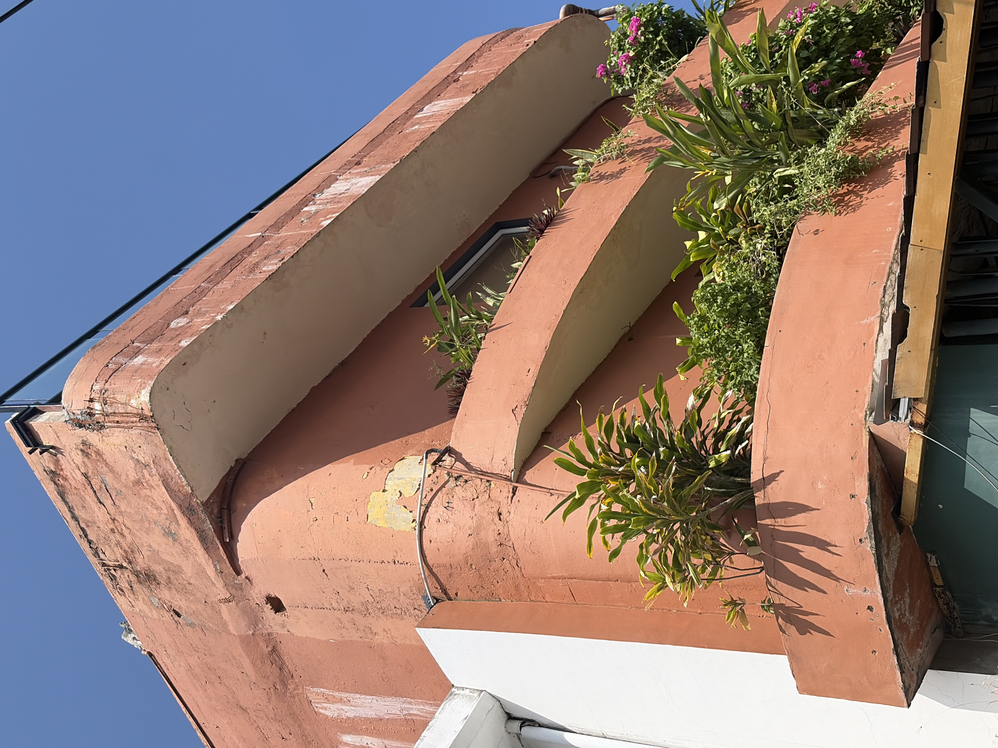 The image shows a close-up view of a weathered, salmon-colored building with multiple balconies filled with lush green plants and vibrant pink flowers. The building's facade exhibits signs of age, with peeling paint and visible cracks. A white column juts out from the left side of the building, contrasting with the dominant pink hue. The sky above is a clear, bright blue. In the foreground, shadows from the plants are cast on the building's surface, indicating a sunny day. The overall impression is one of an urban environment in Kuta, Indonesia, where nature has been integrated onto the architecture, creating a somewhat rustic and tropical aesthetic.