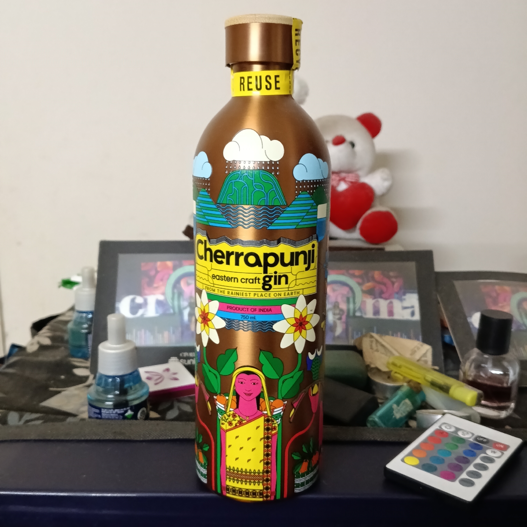 The image depicts a bottle of Cherrapunji eastern craft gin, positioned centrally on a dark blue surface. The bottle has a metallic copper finish and is adorned with colorful illustrations of landscapes, flowers, and a woman in traditional attire carrying baskets. A yellow label with the word "REUSE" is visible on the neck of the bottle. Behind the bottle, on a white wall, sits a white teddy bear with red accents. To the left of the bottle, several small items are visible, including a blue air freshener plug-in and a framed picture. To the right, a small bottle with a black cap containing a dark liquid and a remote control with various colored buttons are present. The overall environment appears to be an indoor setting, possibly a shelf or desk, with the background showing a light-colored wall. There is no indication of the location being specifically in Bengaluru, India; the text on the bottle states "PRODUCT OF INDIA."