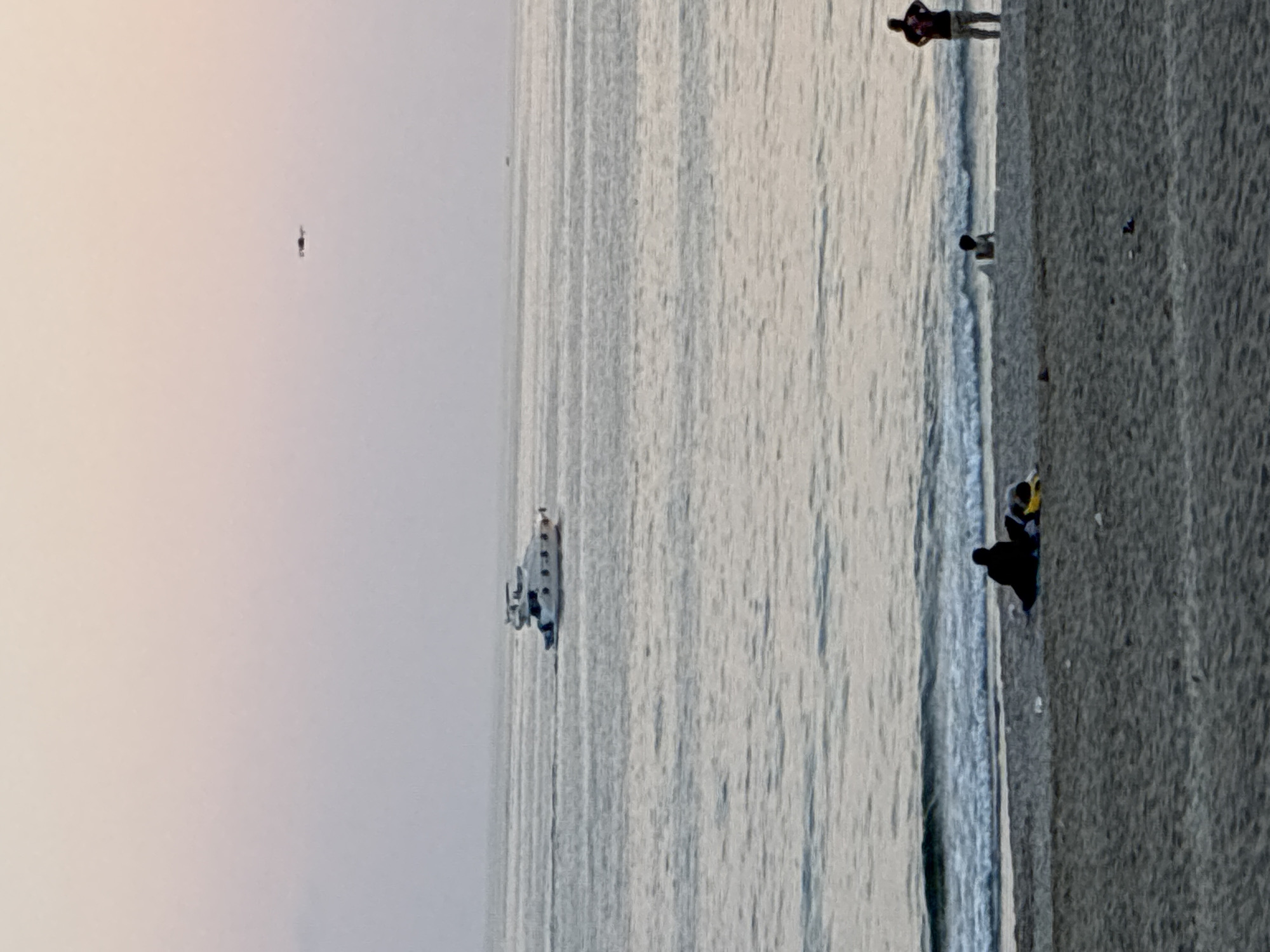 A beach scene at dusk is depicted, with the sky transitioning to soft pastel colors. In the distance, a white yacht is cruising on the calm sea. Closer to the shore, a few individuals are present on the sandy beach. One person is silhouetted on the sand near a yellow object, possibly a bag or blanket. Further to the right, another person stands facing the water, and a third person is partially visible sitting near the water's edge. A small object, potentially a drone, is visible in the sky. The image does not provide specific details to confirm the location in Alanya, Turkey, but it captures a tranquil evening at a coastal environment.