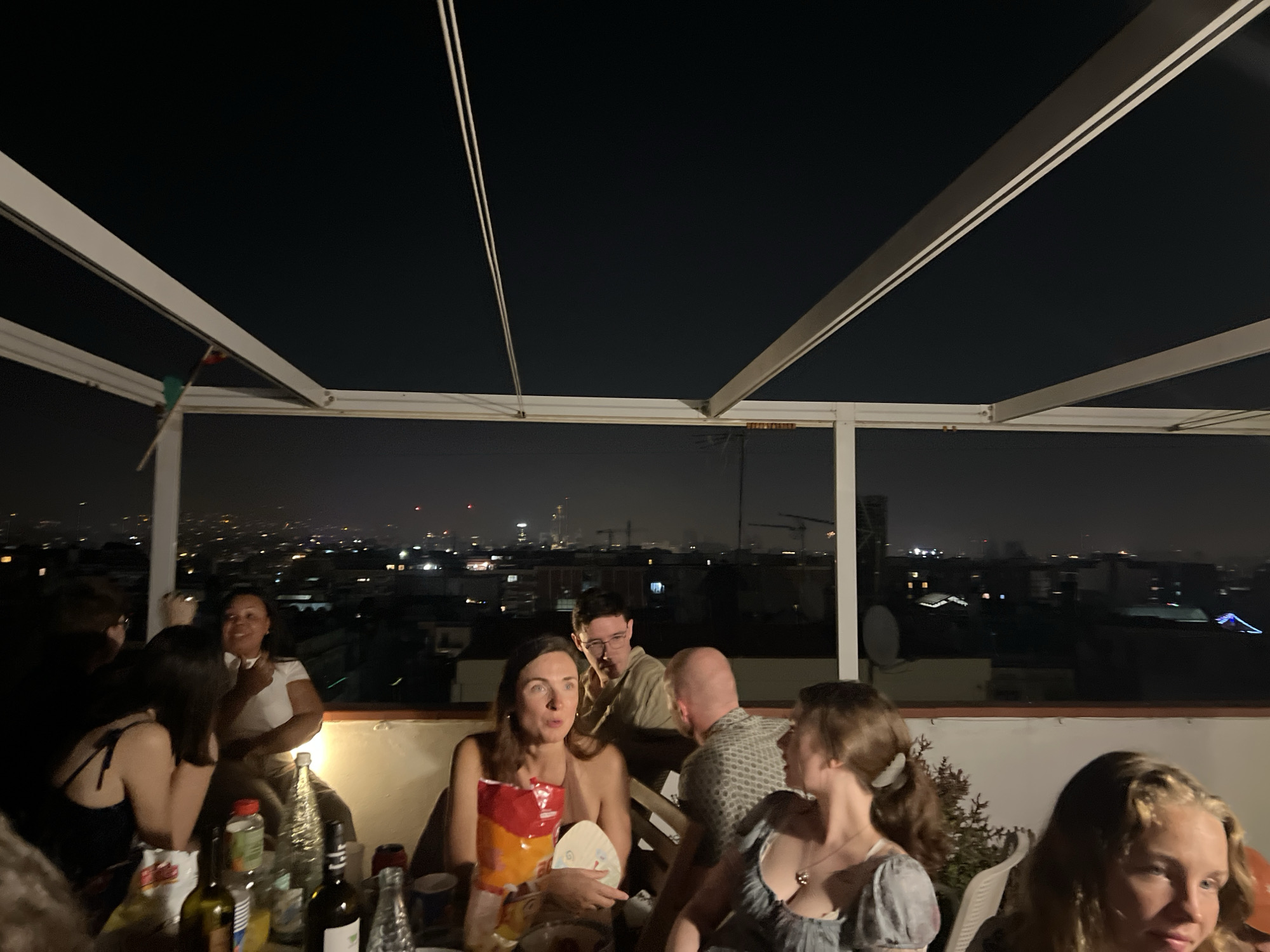 A group of people are gathered on a rooftop terrace at night, overlooking a cityscape that appears to be Barcelona, Spain, with its distinctive architecture and illuminated skyline. The terrace is covered by a white, modern structure with slanted panels, suggestive of an open-air dining or social area.
In the foreground, a table is laden with drinks and snacks. A woman with long brown hair, wearing a beige top, is holding a bag of chips and a white plate. To her right, a woman with blonde hair, wearing a grey top, is also present. Behind them, a man with glasses and a light-colored shirt is seated, looking towards the right. To his left, another man with a patterned shirt is visible.
Further back, on the left side of the image, a woman in a white top and light-colored pants is seated, gesturing with her right hand. She appears to be mid-conversation. To her left, a woman with dark hair is seated.
The lighting is dim, indicative of nighttime, with artificial lights from the city below providing illumination. The overall atmosphere suggests a social gathering or party.