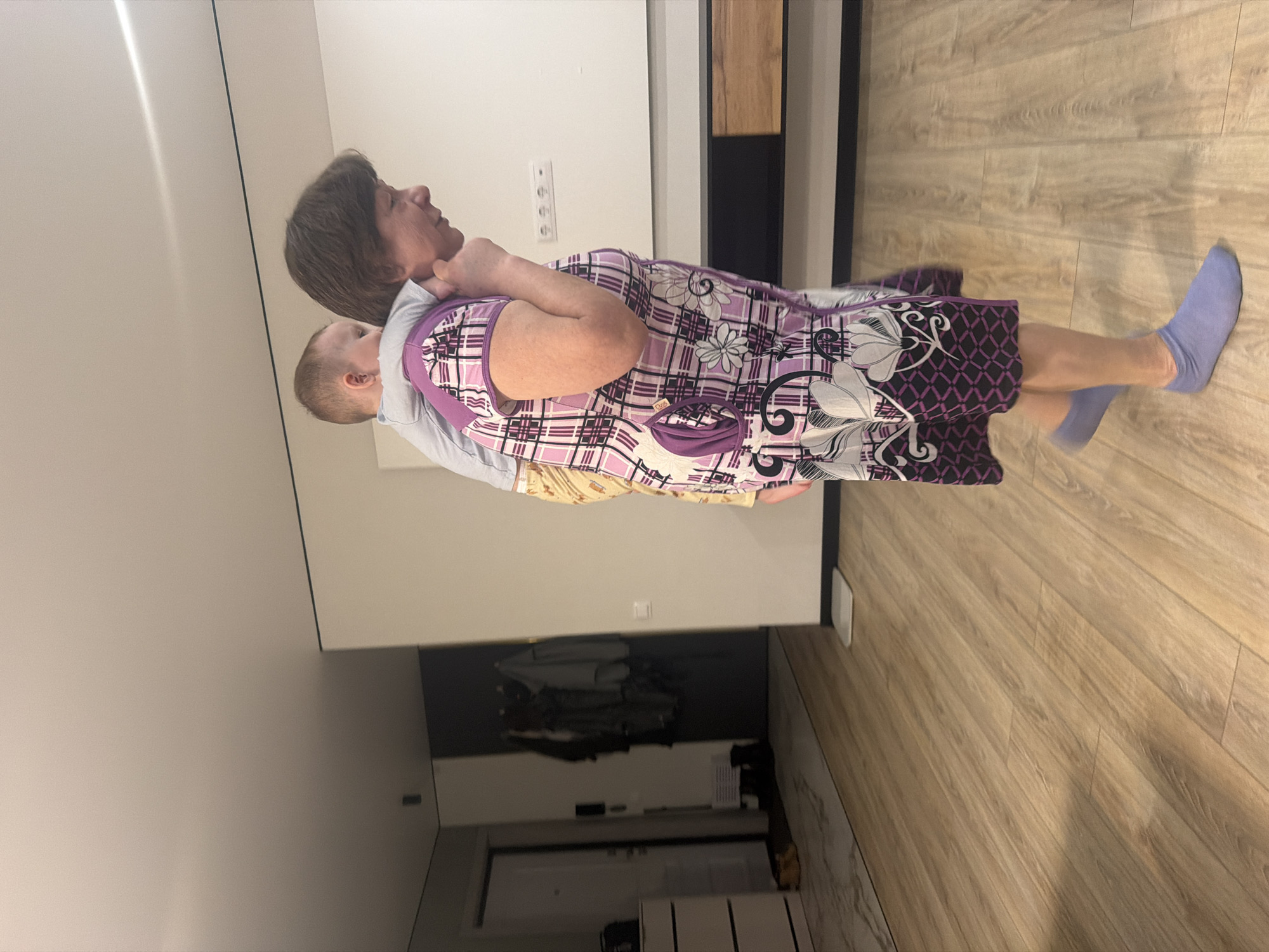 A woman with short brown hair is standing in an indoor room, carrying a baby on her back using a carrier. The woman is wearing a long, patterned dress and blue socks. The room has light-colored wood floors and white walls. The location is unknown, but the possibility of Chaikovsky, Russia is offered. A cabinet and doorway are visible in the background.