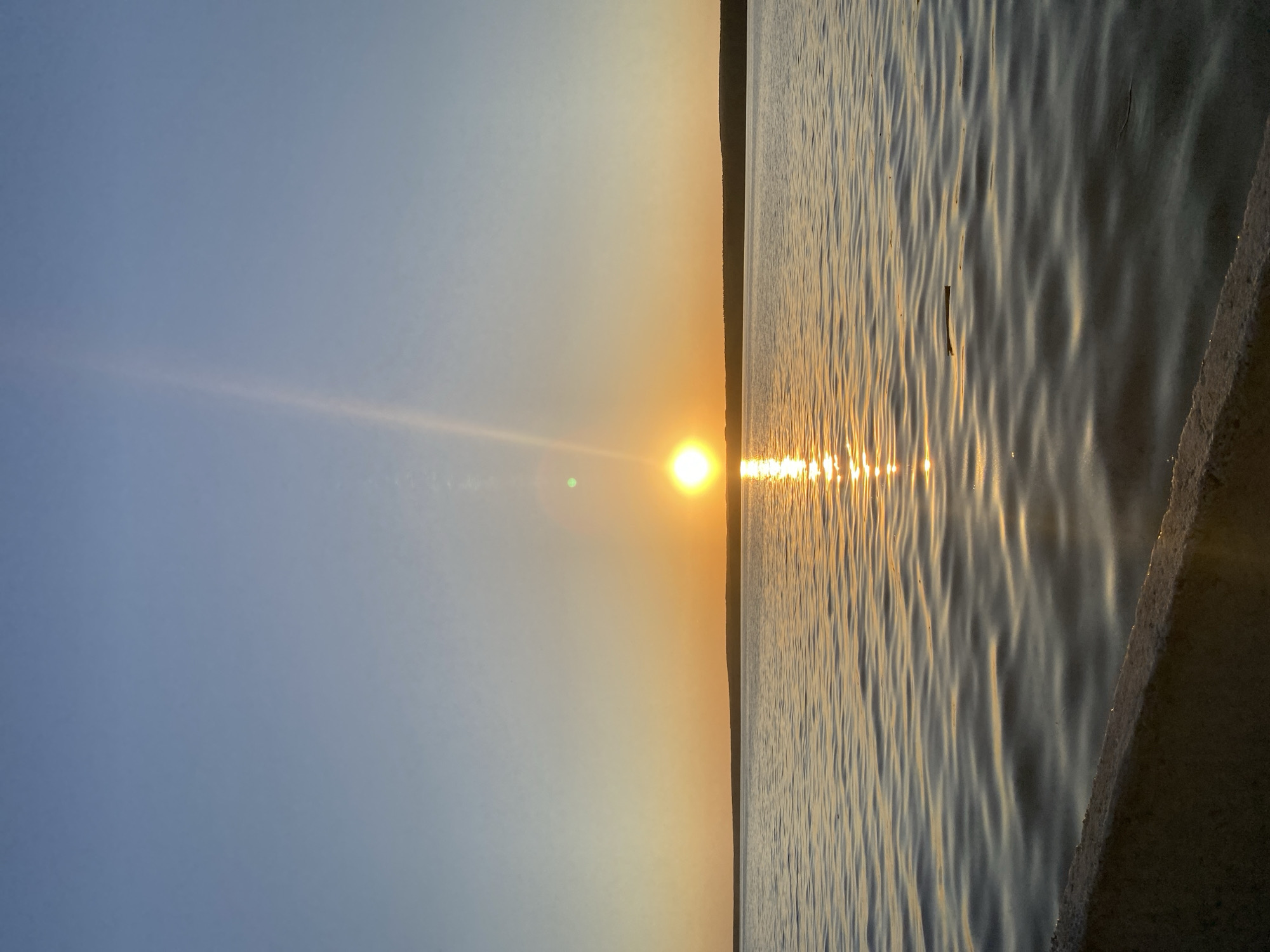 The image shows a sunset over a body of water, likely a lake or river. The sun is low on the horizon, casting a bright reflection on the water's surface. The sky is clear with gradations of blue to orange near the horizon. A faint line of trees or land is visible on the horizon between the sky and water. The foreground includes a concrete structure, possibly a bank or dock. The location is in the city of Chaikovsky, Russia. The water surface is rippled. A stick or piece of debris floats on the surface.
