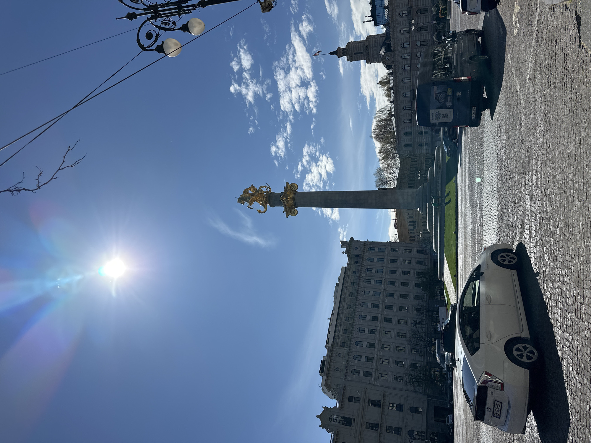 The image shows the Freedom Monument in Tbilisi, Georgia, under a bright blue sky with scattered clouds. The monument, topped with a golden statue of St. George slaying a dragon, stands prominently in the center. Various buildings surround the monument, including a structure with a clock tower visible in the background. The streets are paved with cobblestones, and there is a white car parked to the right and a black van parked farther. A decorative lamppost is visible. The sun is shining brightly, creating a lens flare.
