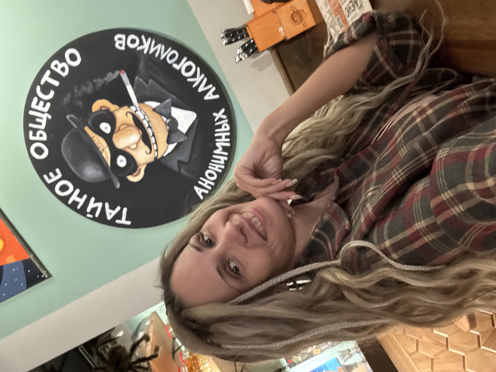 The image captures a young woman smiling and posing indoors, possibly within a cafe or themed establishment in Rusanovo, Russia. She has long, wavy blonde hair with several braided strands and is wearing a plaid shirt in shades of red, green, and dark grey. Her head is tilted slightly, and her right hand is near her chin as she looks cheerfully at the camera.
Behind her, mounted on a light green wall, is a prominent large, circular black sign. The sign features a cartoon caricature of a man in a bowler hat, suit, and bow tie, sporting a large nose, round glasses, and a small mustache. He is depicted smoking a cigarette, with white smoke emanating from it. White capital letters are arranged in a circle around the character. The text reads: "ТАЙНОЕ ОБЩЕСТВО АЛКОГОЛИКОВ АНОНИМНЫХ," which translates to "SECRET SOCIETY OF ANONYMOUS ALCOHOLICS."
The background also shows glimpses of a wooden table with a hexagonal pattern in the lower right, and some orange boxes and what appear to be cutlery holders in the upper right. The setting suggests an indoor environment, potentially a casual dining or social space. The woman's expression conveys a friendly and relaxed mood.