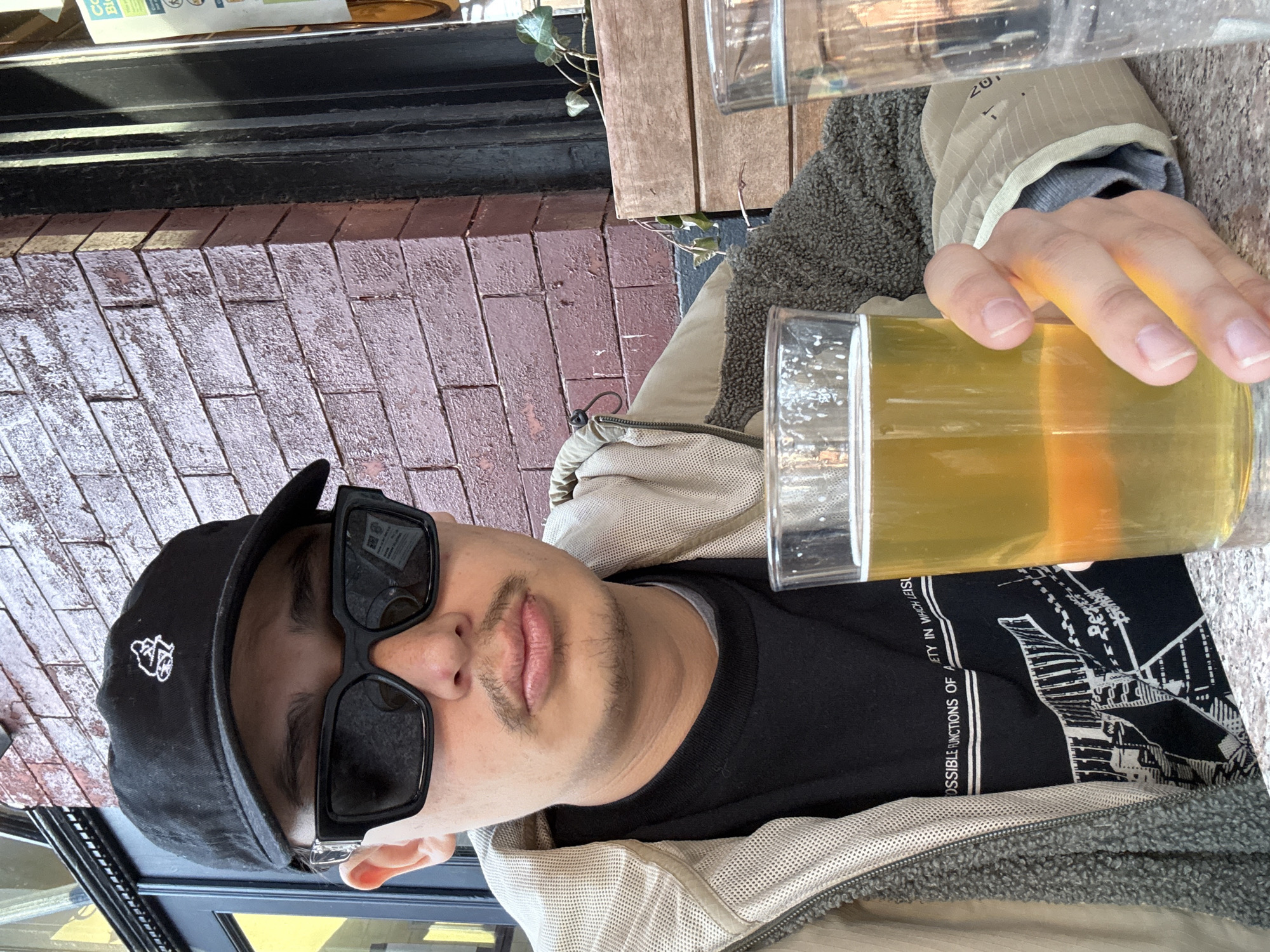 A person is seated outdoors, holding a glass of beer. The individual is wearing a black baseball cap with a white logo, large dark sunglasses, and a black T-shirt. A light-colored jacket with a gray fleece lining is visible. The person's hand is touching the side of the beer glass. Part of a building with red brickwork and a black window frame is in the background. The image suggests the person may be at a restaurant or cafe in an urban area. The text on the black T-shirt is visible, it includes the phrase "POSSIBLE FUNCTIONS OF AETY IN WHICH LEISU". It's not possible to determine the exact location in New York City from the given image.