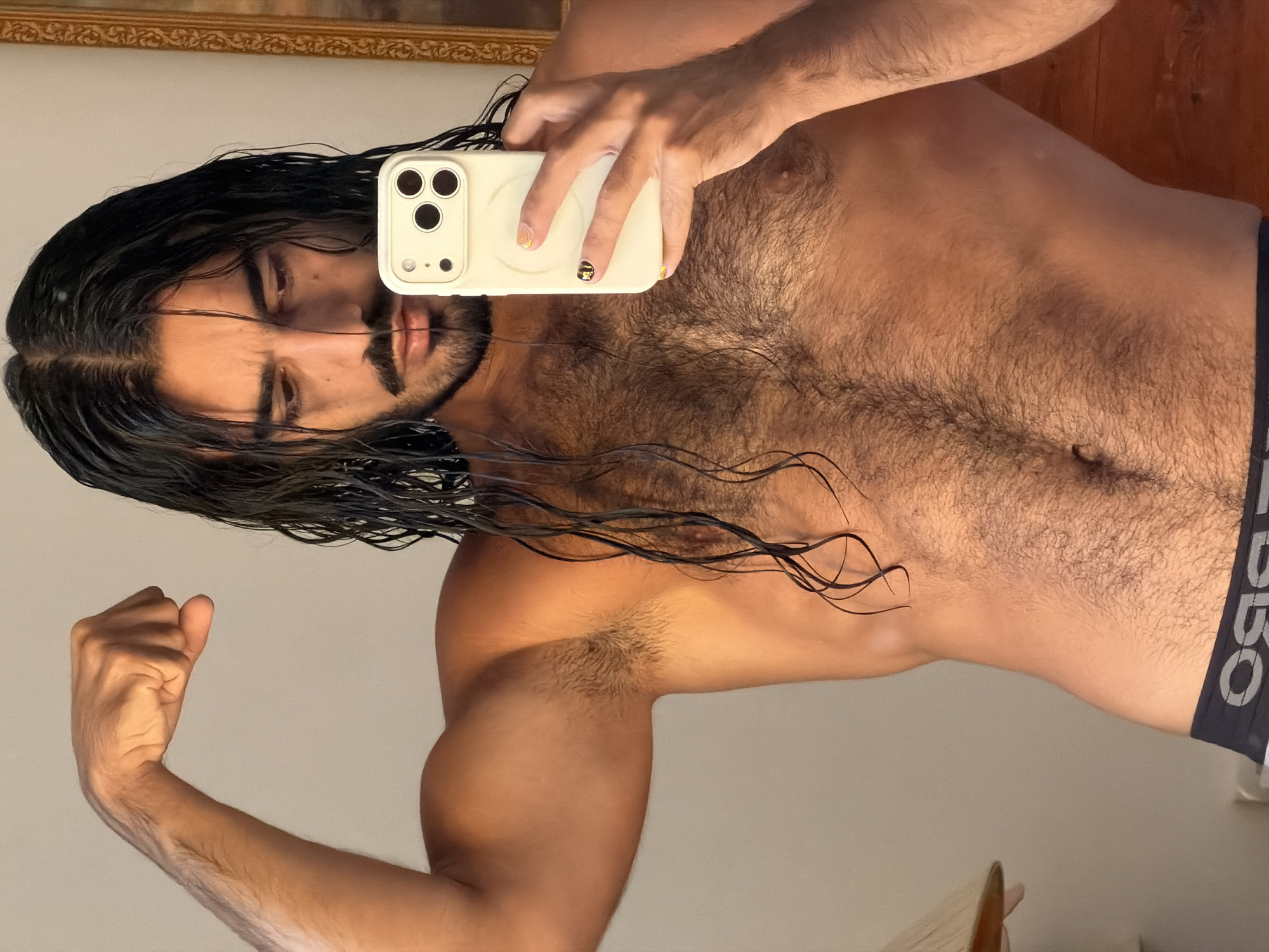 In North Kuta, Indonesia, a man with long, dark, wet hair and a mustache is taking a selfie in a mirror. He is shirtless, flexing his bicep and showing his hairy chest. He is holding a white iPhone in his right hand. He is wearing dark underwear. The background is a plain wall with a framed picture on the right.