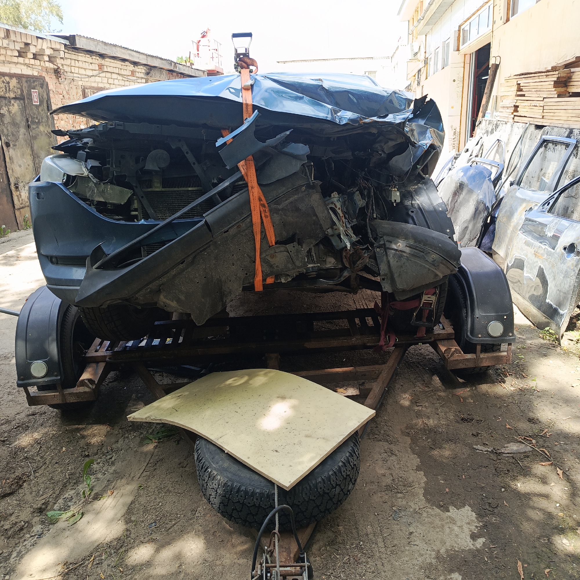 A severely damaged blue car, its front end crushed and buckled, is secured to a trailer with orange straps. The car is positioned on a metal trailer with two wheels and fenders. In the foreground, a spare tire sits on the trailer, covered by a light-colored rectangular board. Behind the trailer, several car doors, some showing signs of damage and rust, are leaning against a wall. The environment appears to be an outdoor industrial or junkyard area, with a brick building and wooden structures visible in the background. The ground is dirt and gravel, with some scattered leaves. The lighting suggests it is daytime. The location is identified as Chaikovsky, Russia. There are no people visible in the image.
