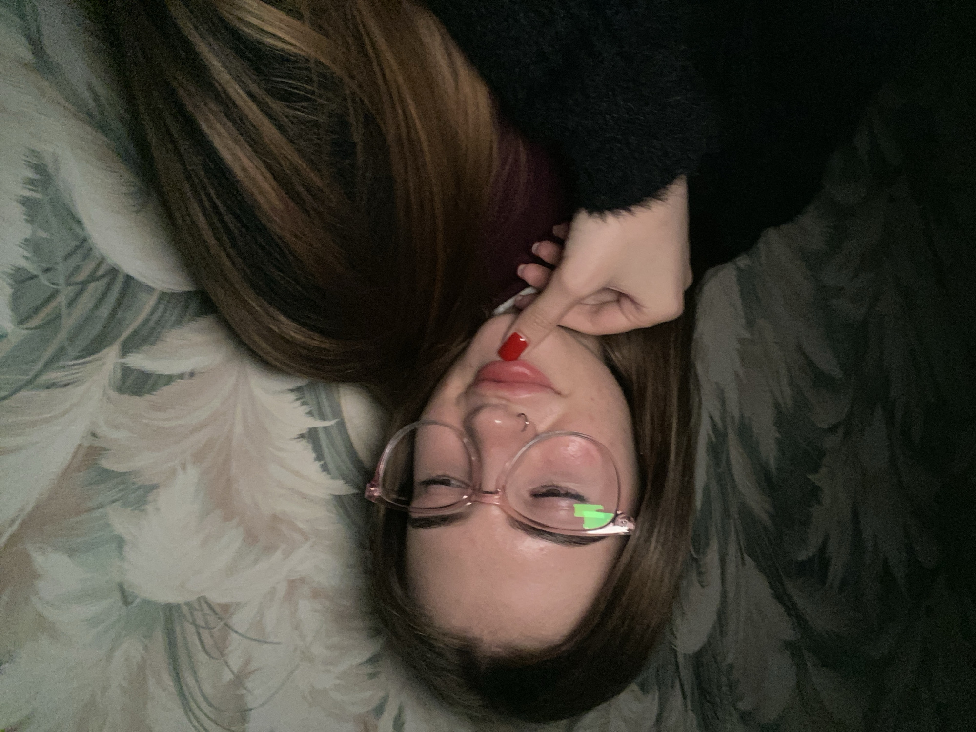 The image captures a close-up, high-angle view of a person lying down, facing upwards towards the camera. The individual, likely female, has long, straight, brown hair that frames her face. She is wearing light pink-framed glasses, through which her eyes appear mostly closed or squinted. A small silver hoop piercing is visible on her left nostril. Her lips are slightly parted, showing their natural light pink hue.
Her left hand, with a distinctive bright red nail polish on the index finger, is gently positioned near her upper lip. She is dressed in a dark, fuzzy garment, possibly a sweater. The background is a patterned fabric, resembling a bedspread or upholstery, featuring light, feathery or floral designs on a muted blue-green and dark grey base. The lighting is soft and appears to be indoors. A distinct, bright green rectangular reflection is visible on the right lens of her glasses. No text is visible in the image. The scene conveys a relaxed and intimate atmosphere, possibly a moment of rest or a casual selfie.