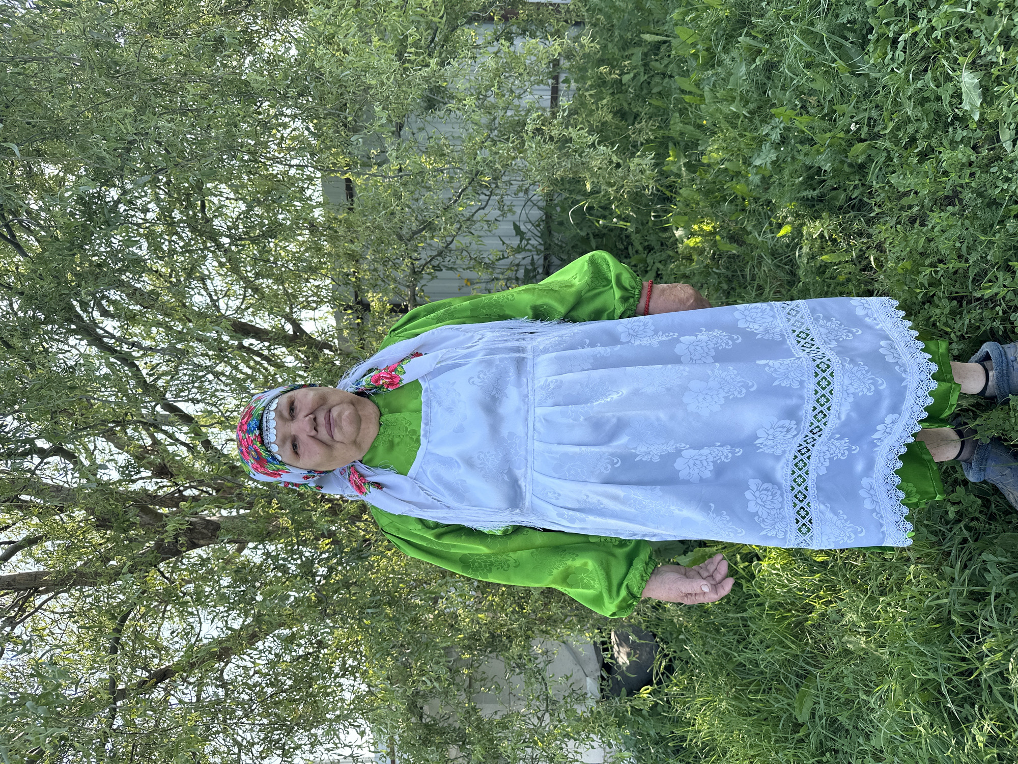 An elderly woman stands outdoors in what appears to be a village setting, possibly in Sredneye Kechevo, Russia. She is positioned in front of a large, leafy tree and a light-colored fence. The woman is wearing a vibrant green, long-sleeved blouse with patterned fabric and a white apron over it. The apron is decorated with floral embroidery and lace trim. She also wears a colorful headscarf with floral patterns and a white kerchief tied around her neck. Her feet are clad in dark, practical footwear. The surrounding environment is overgrown with green grass and various plants, suggesting a rural or natural setting. The lighting indicates it is daytime, with sunlight filtering through the tree branches.