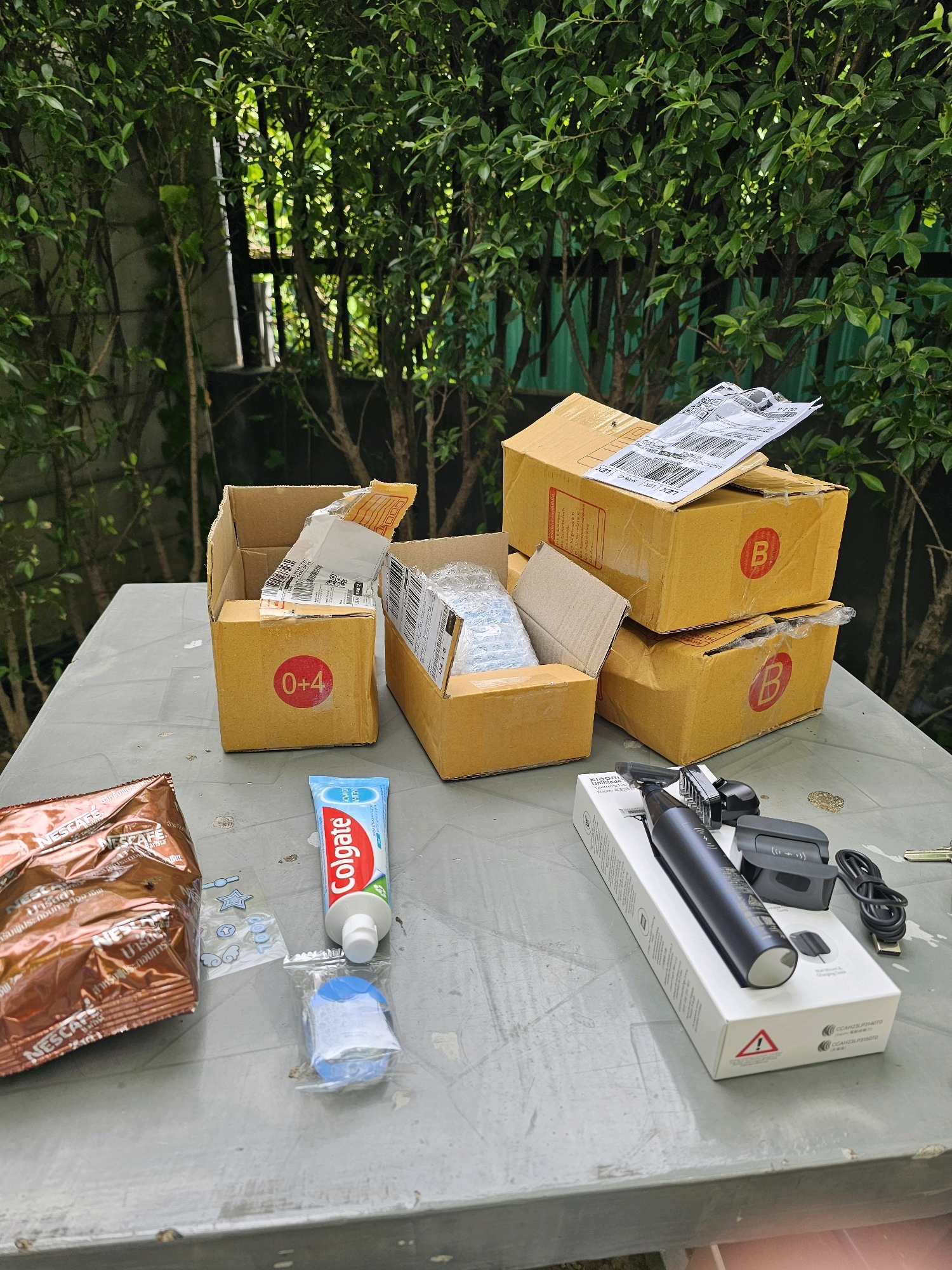 The image depicts a table outdoors, likely in a residential area of Bangkok, Thailand, with various items placed on it. The background features dense green foliage and a fence.
On the table, there are several cardboard boxes. Two are stacked on top of each other, both brown with red circular markings containing the letter 'B'. Another brown cardboard box is open, revealing some packing material and shipping labels. A smaller open box, also brown, has a red circular sticker with "0+4" written on it.
In the foreground, several consumer products are visible. A brown packet of Nescafe coffee is on the left. Next to it is a tube of Colgate toothpaste. To the right of the toothpaste is a small, clear plastic package containing a blue object. Further to the right, a white box displays a black electric trimmer, a charging stand, and a USB cable. A small gold-colored item, possibly a coin or a small accessory, is also present. The table itself appears to be made of a gray, textured material. The overall scene suggests unpacked items, possibly from a delivery.