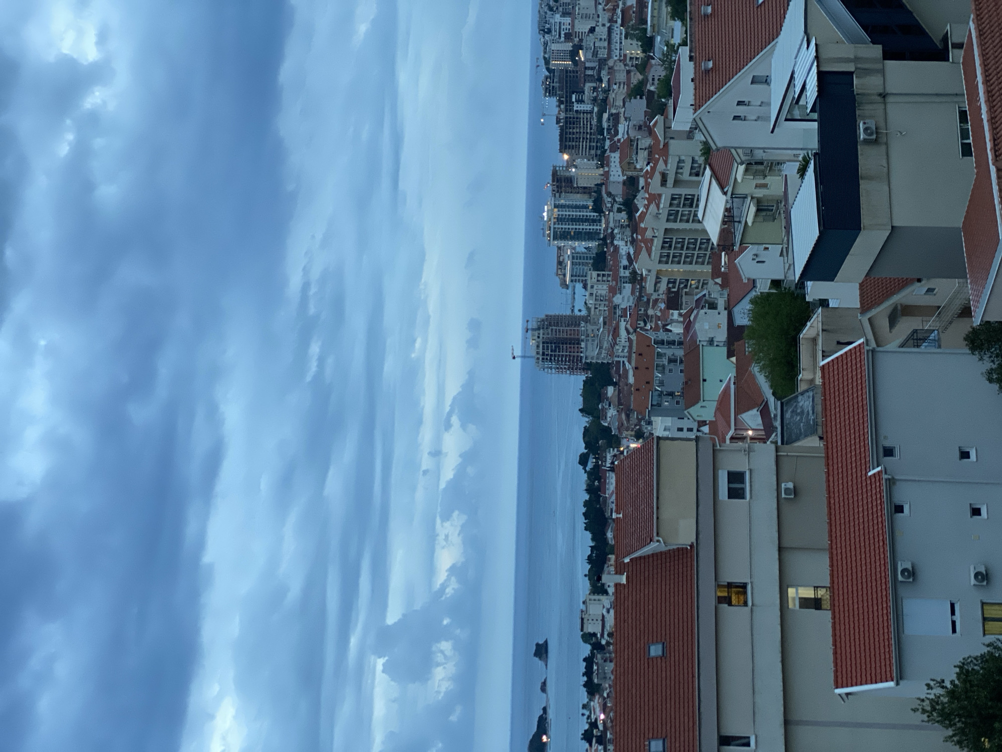 The image displays a panoramic view of a coastal city, identified as Budva, Montenegro, observed from an elevated vantage point. The scene depicts a twilight hour or a heavily overcast day, characterized by a muted color palette dominated by blues, grays, and terracotta.
The upper portion of the frame is filled with a vast, cloudy sky. Dense, elongated grey-blue clouds extend across the expanse, creating a dramatic and somber atmosphere. Below the sky, the calm, grey-blue water of the sea stretches horizontally to meet the distant horizon. Several small, dark islands or landmasses are visible on the water's surface in the midground.
The lower half of the image is occupied by the city's architecture. In the foreground and midground, numerous buildings with characteristic terracotta-red tiled roofs are prominent, indicative of Mediterranean design. These structures appear to be a mix of residential and commercial properties. Many windows are visible, with some illuminated, suggesting evening time or interior lights on during a dark day. Further along the coastline and receding into the background, taller, more modern multi-story buildings, likely apartment blocks or hotels, punctuate the skyline. Some construction activity, including cranes, can be discerned amidst the urban development in the distance. Sparse green trees and foliage are scattered throughout the cityscape.
No people or specific activities are visible, and there is no discernible text within the image. The overall impression is one of a serene yet densely populated coastal settlement under a dramatic sky.