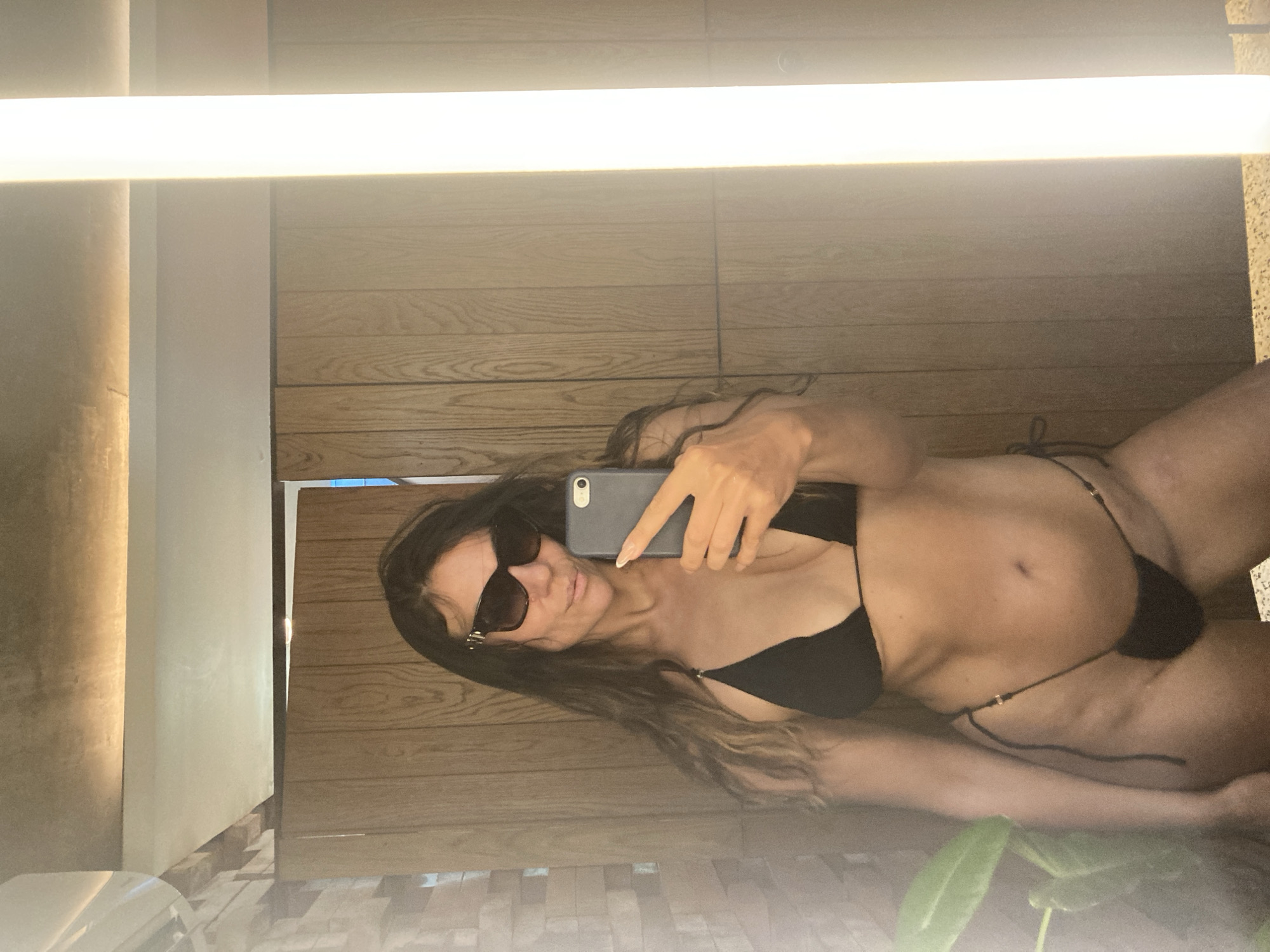 A woman takes a mirror selfie in a bathroom with wooden paneling and a bright overhead light. She is wearing a black bikini and sunglasses, and is holding her phone to take the picture. The setting appears to be indoors, possibly a modern spa or hotel bathroom given the materials and lighting. The lighting is bright and even, suggesting it could be daytime or well-lit at night. No specific time of day or weather conditions are discernible. The woman's expression is neutral as she focuses on taking the photo. There is no visible text in the image.