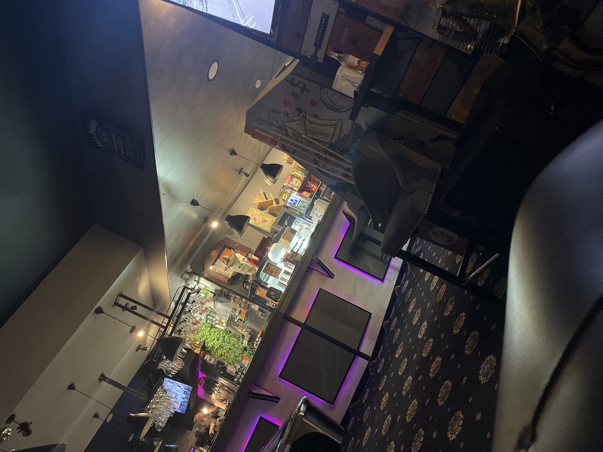 The image depicts the interior of a bar/restaurant, possibly in Plovdiv, Bulgaria, though this cannot be verified solely from the image. The scene features a bar area with shelves containing bottles, glassware, and greenery. Several pendant lights illuminate the bar, and there is a television screen in the upper portion of the frame. The flooring has a patterned design. A bar stool is situated near the bar. There are no readily visible people engaging in specific actions or interactions.