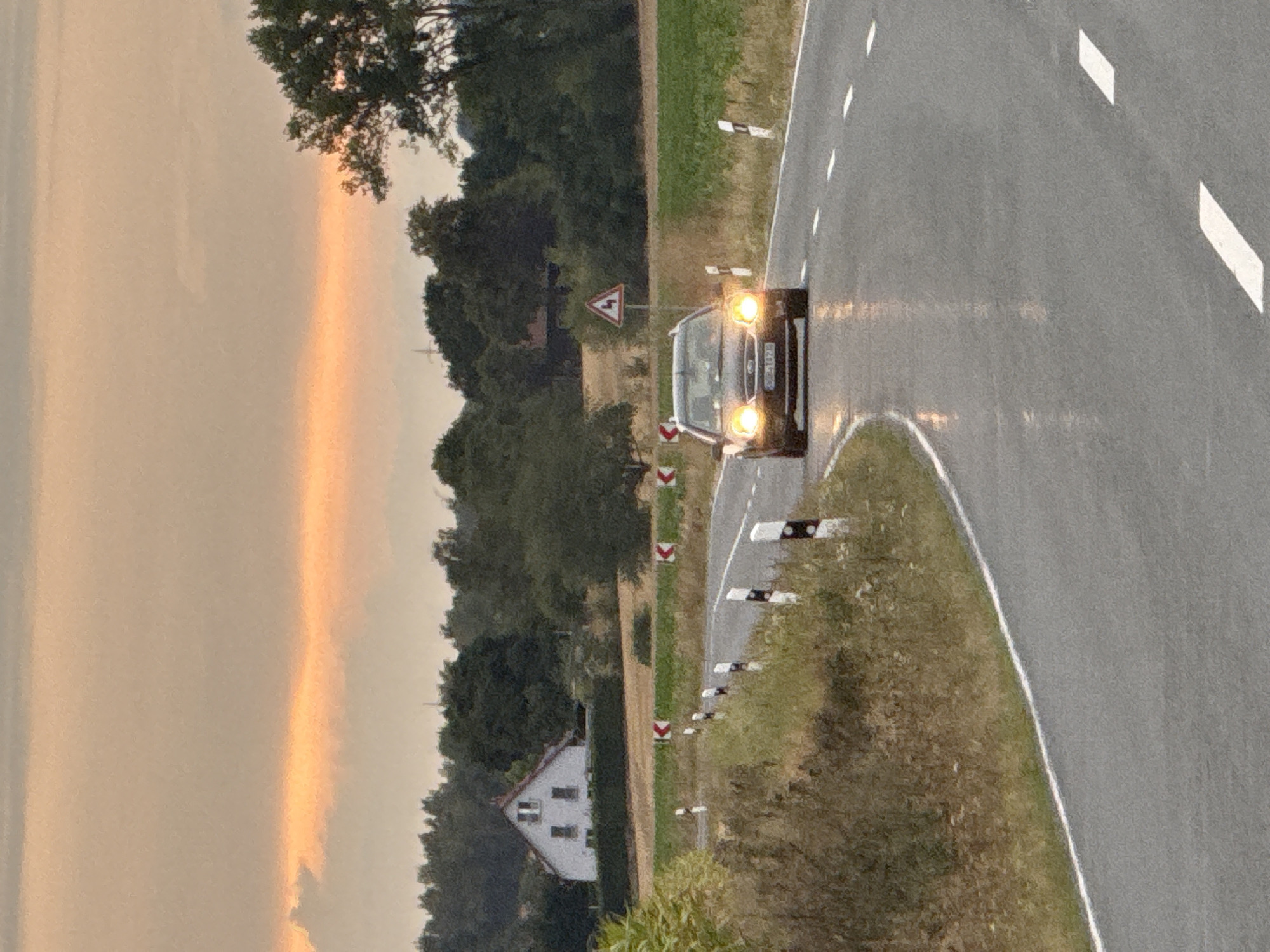 The image shows a road in the rural area surrounding Hohenstein, Germany. The road curves to the right and appears wet, with reflections of the car's headlights on its surface. A dark-colored car is driving on the road with its headlights on. The car has a license plate that is partially visible. On the left side of the road, there is a grassy embankment with some dry vegetation. A row of black and white bollards with reflective markers lines the side of the road, indicating a curve. In the background, there is a white house with a red roof, surrounded by trees. Further back, there is a dense line of trees and a field of crops. The sky is overcast with a soft orange glow near the horizon, suggesting either sunrise or sunset. There is a triangular road sign with a red border and black arrows indicating a series of curves. The scene is captured during daylight hours, likely in the late afternoon or early evening.
